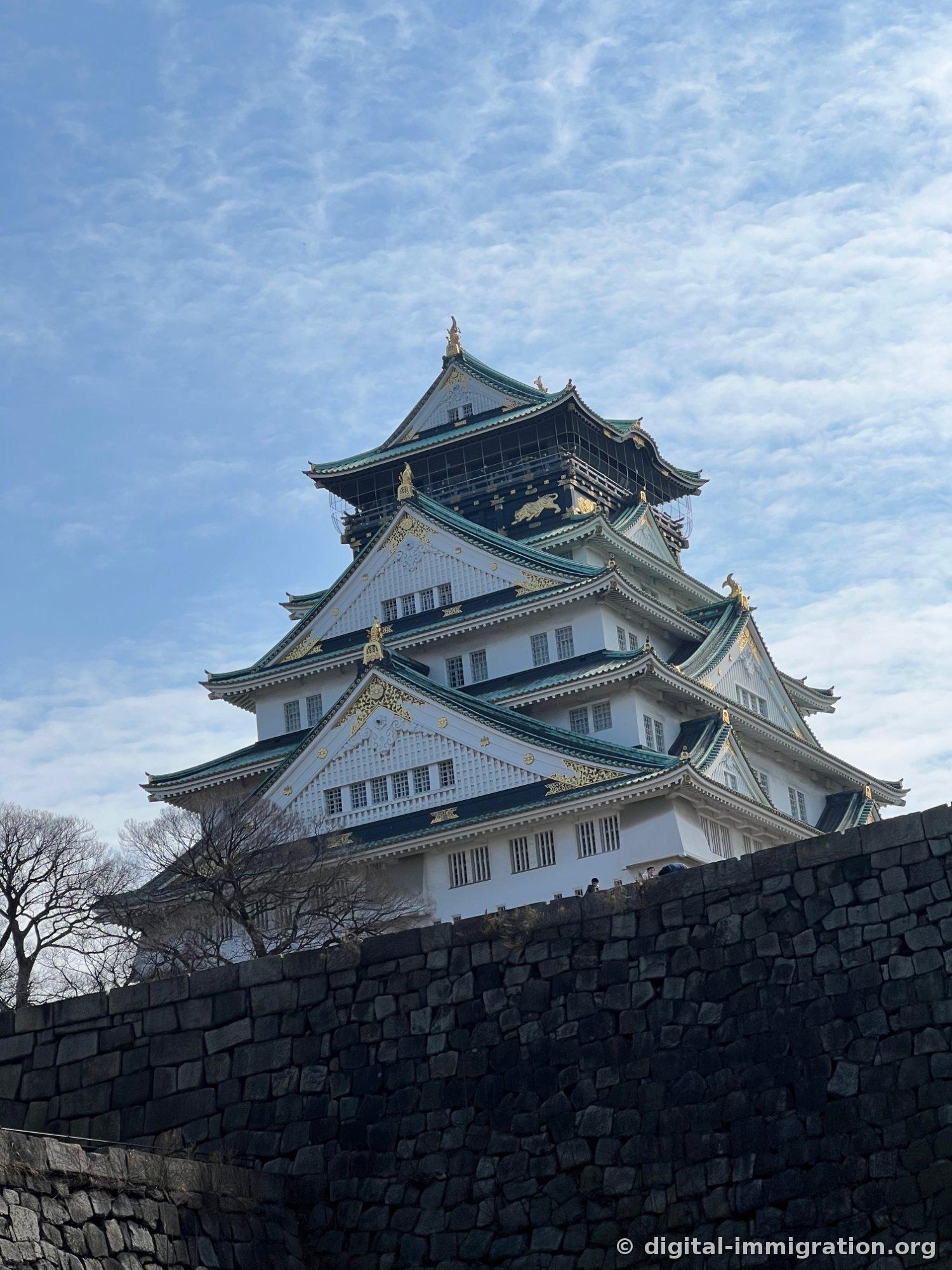 Vue complète de la tour de guet du château d'Osaka debout sur le mur de pierre sous un ciel d'après-midi dégagé. Le mur blanc, les tuiles vertes et les ornements dorés sont clairs