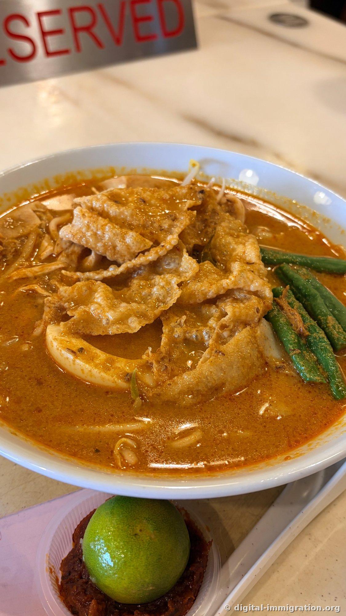 In an orange coconut curry broth sit crispy fried skin, squid, long beans, and bean sprouts in a curry mee bowl, with a sambal dish topped with a lime half in front, and a RESERVED sign in the back
