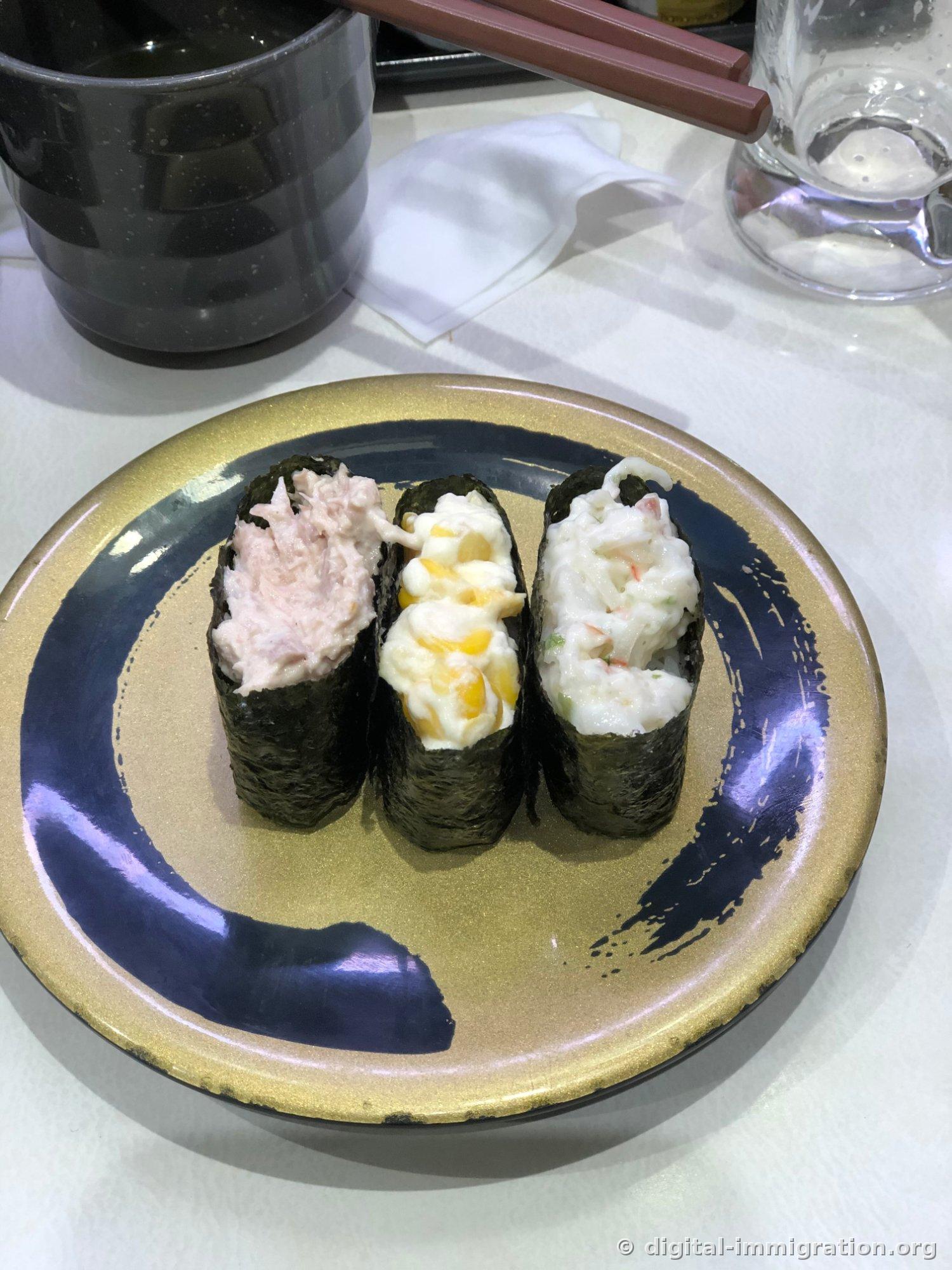 Sur une assiette dorée, trois temaki encerclés d'algue - mayo-thon, mayo-maïs, et salade de crabe - sont disposés côte à côte, avec des baguettes et un verre d'eau à côté