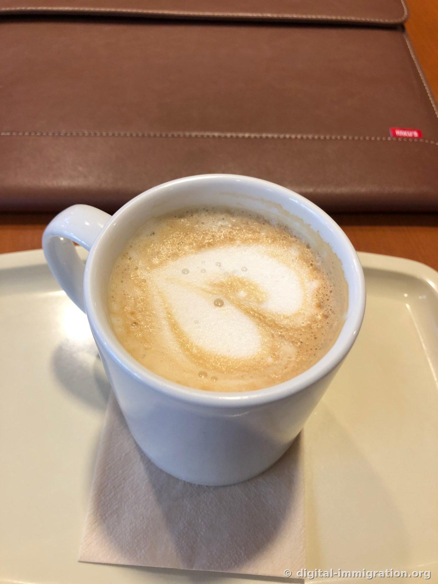 Un café au lait dans une tasse en céramique bleu clair. La mousse de lait s'étend en motif tourbillonnant, posée sur une serviette sur un plateau en plastique ivoire avec un canapé en cuir marron visible derrière
