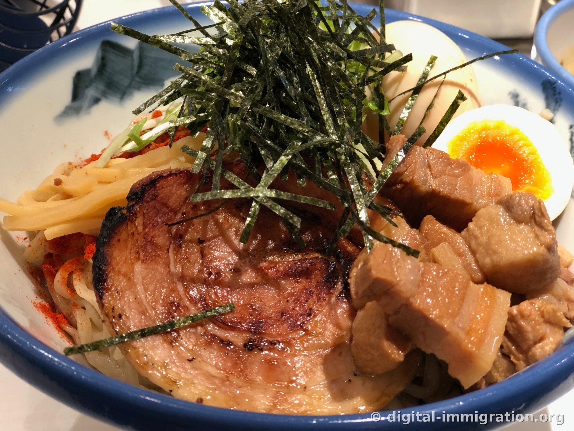 Thick noodles topped with piled shredded seaweed, thick torched chashu, diced braised meat, and half a soft-boiled egg in a blue-rimmed bowl