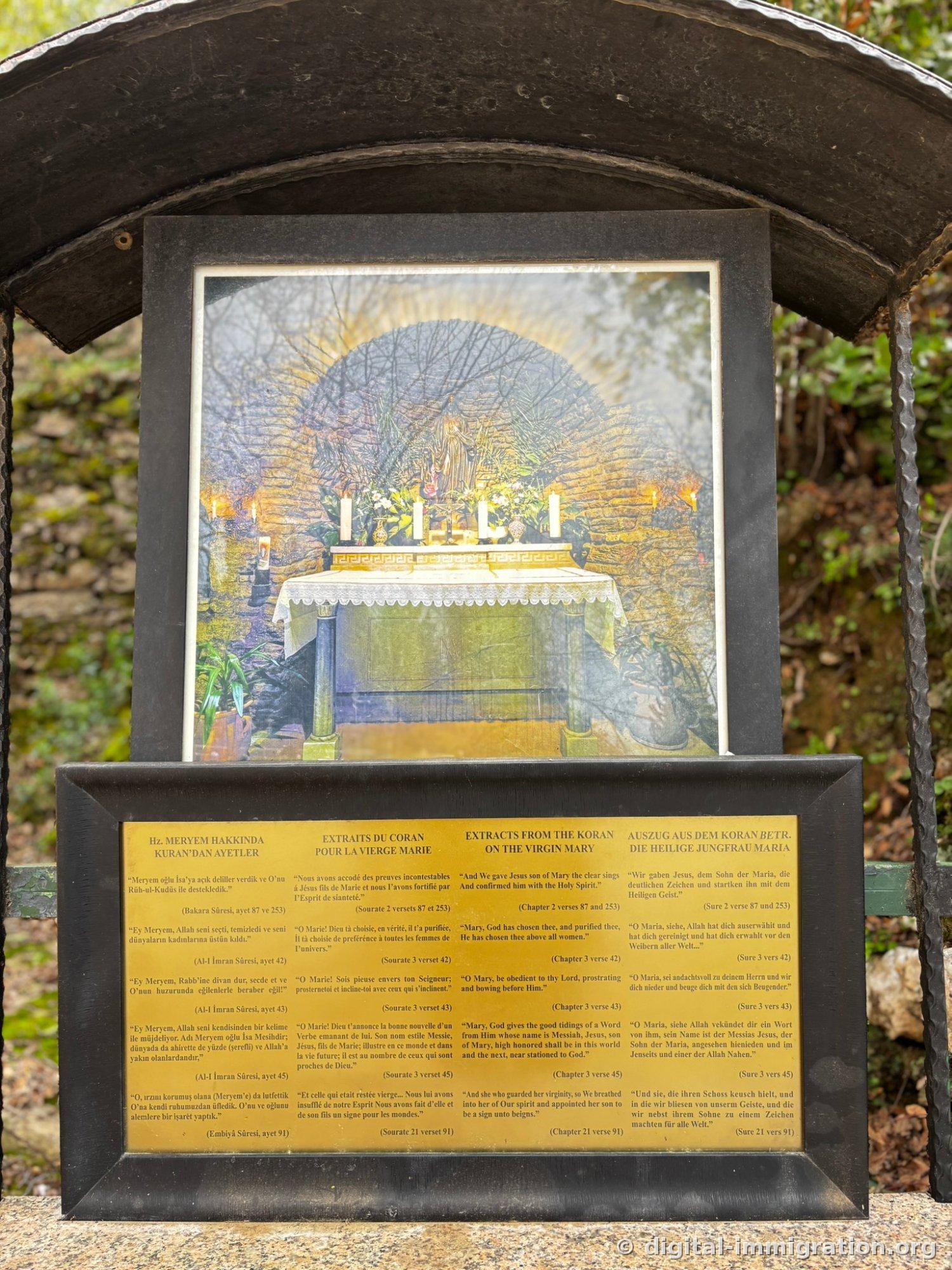 A frame hanging in a display case under a black arch, with a photo of the candlelit altar above and a brass plaque below engraved with Quranic verses about Mary in Turkish, French, English, and German