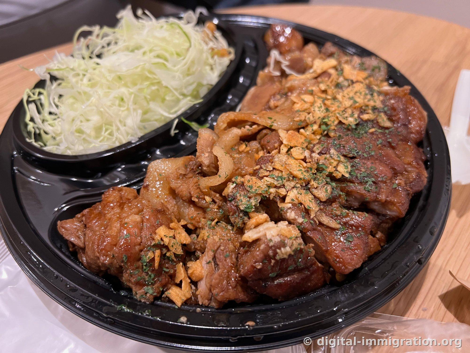 A closeup of pork steak in a partitioned black container. Soy-glazed meat topped with golden fried garlic chips and parsley powder, with finely shredded fresh cabbage in the upper left section.