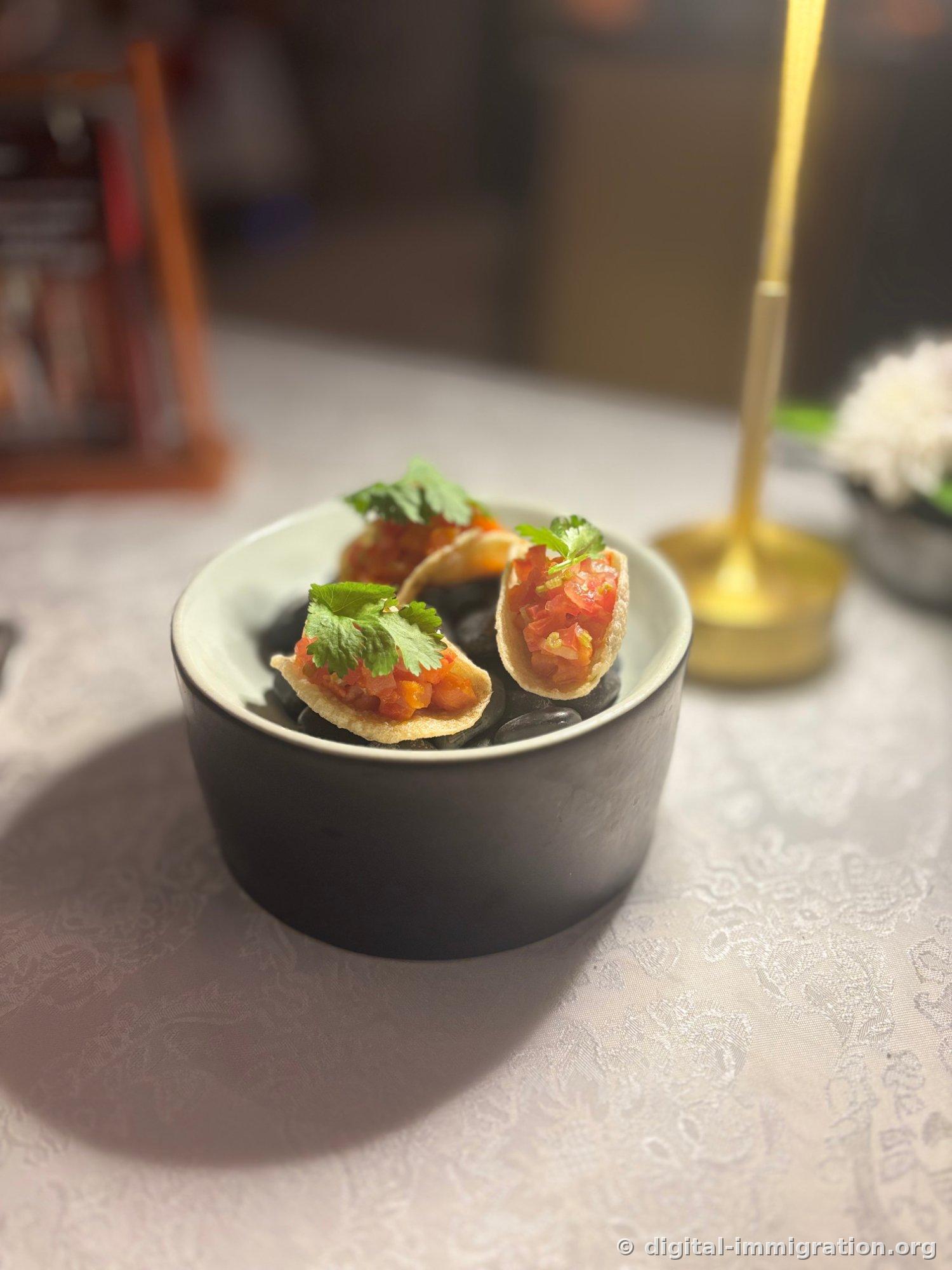 A single-bite appetizer with minced tomato salsa and cilantro leaves on a crispy shell, served on black gravel-lined plate