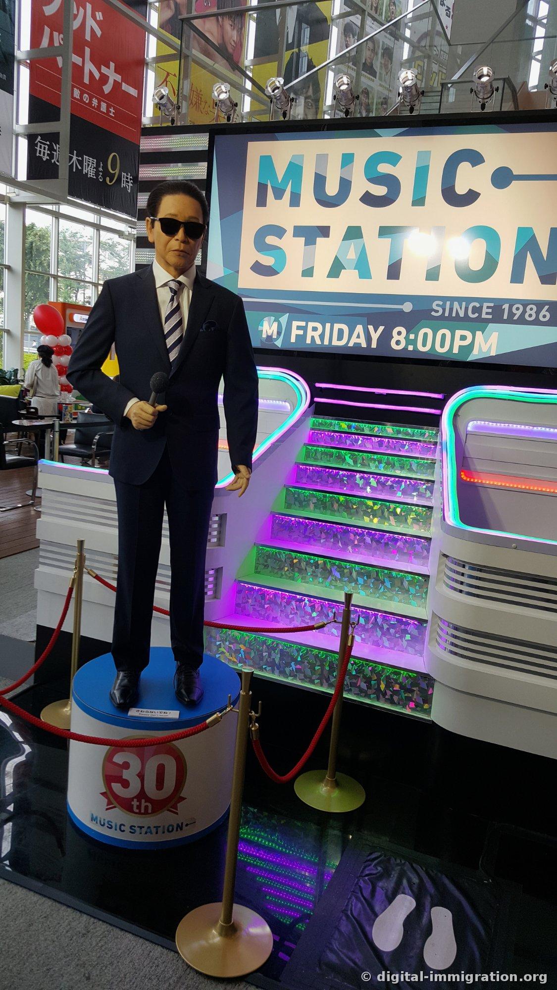 Exposition du 30e anniversaire de MUSIC STATION avec une figurine grandeur nature d'un présentateur en costume tenant un micro et un escalier arc-en-ciel illuminé par des LED à l'arrière, avec l'inscription SINCE 1986 FRIDAY 8:00PM sur l'écran
