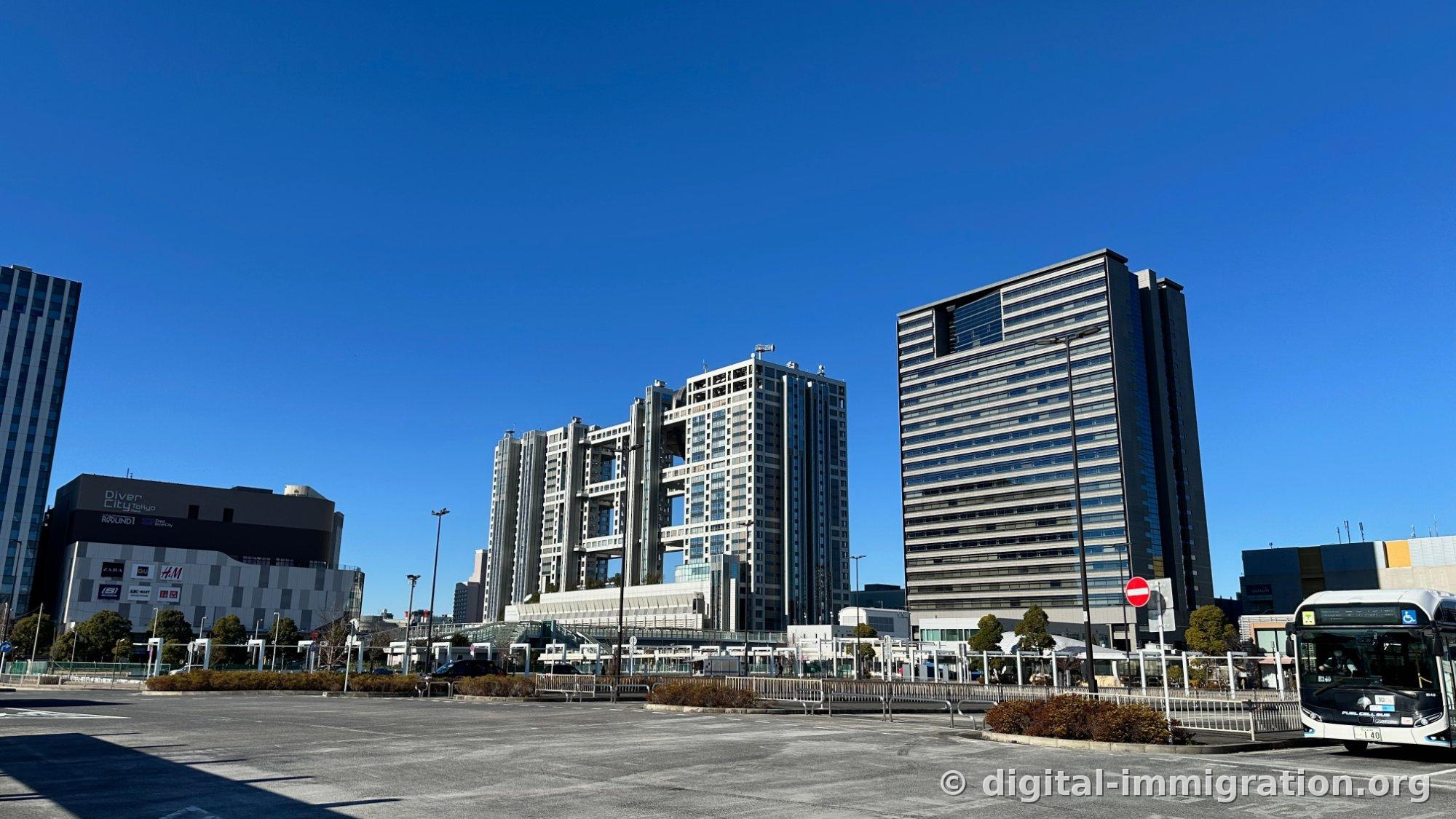 Odaiba street view with the DiverCity Tokyo Plaza building on the left displaying black signage and ZARA·GU·H&M logos under blue sky, Fuji TV headquarters and high-rise buildings visible on the right
