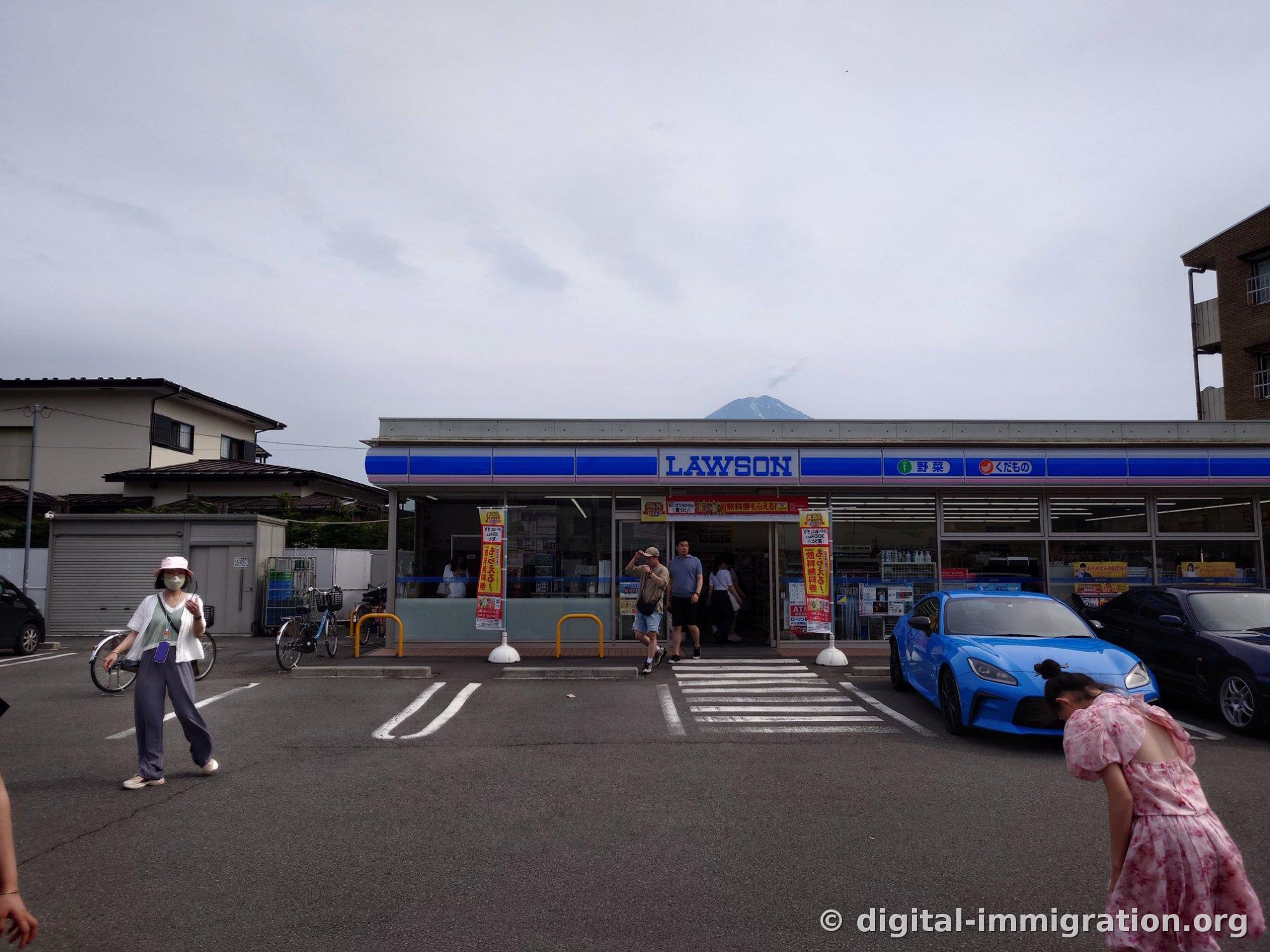 Mount Fuji's peak appears slightly above the blue LAWSON sign, with a blue sports car in the parking lot and tourists moving about