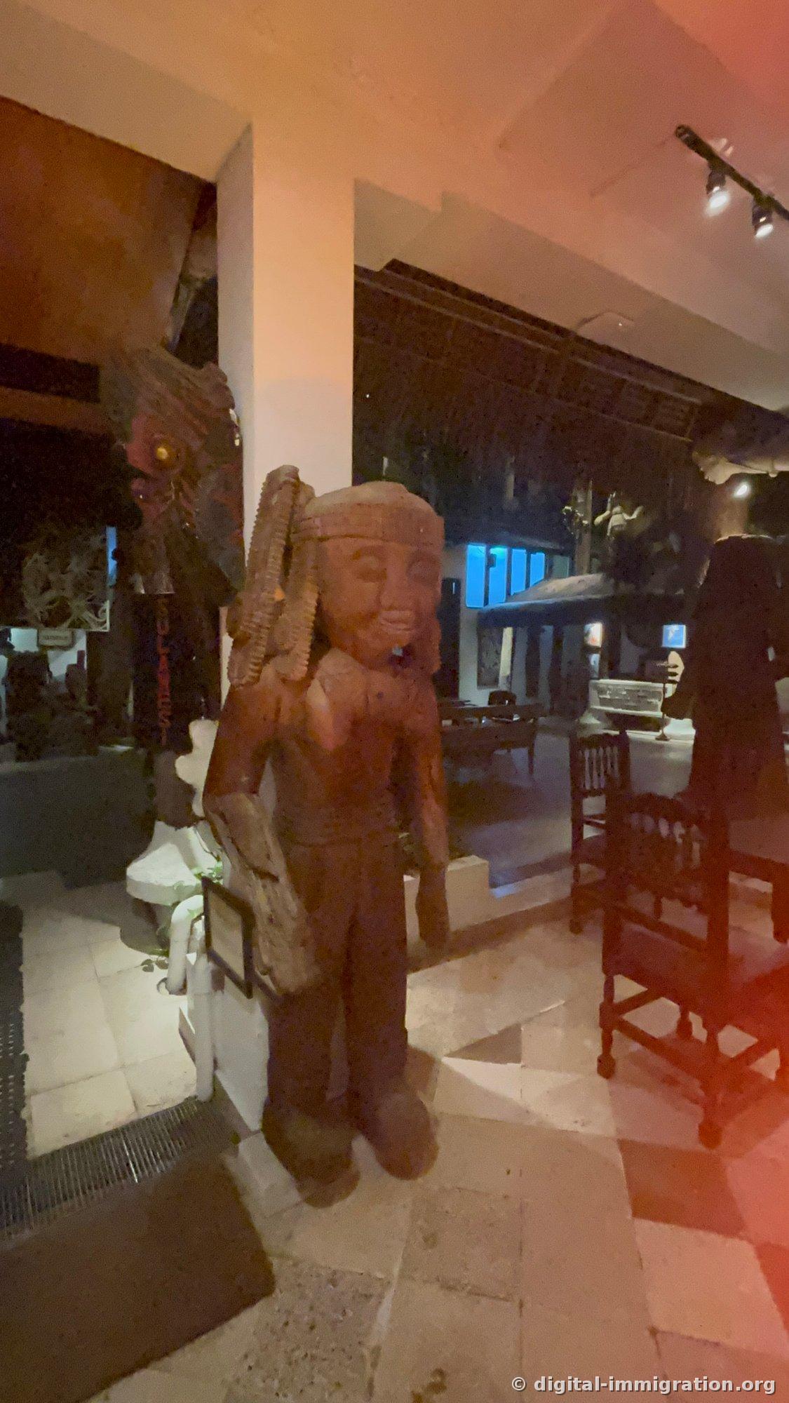Wooden human figure sculpture standing in a corridor with red lighting, posed as if carrying a load on its back