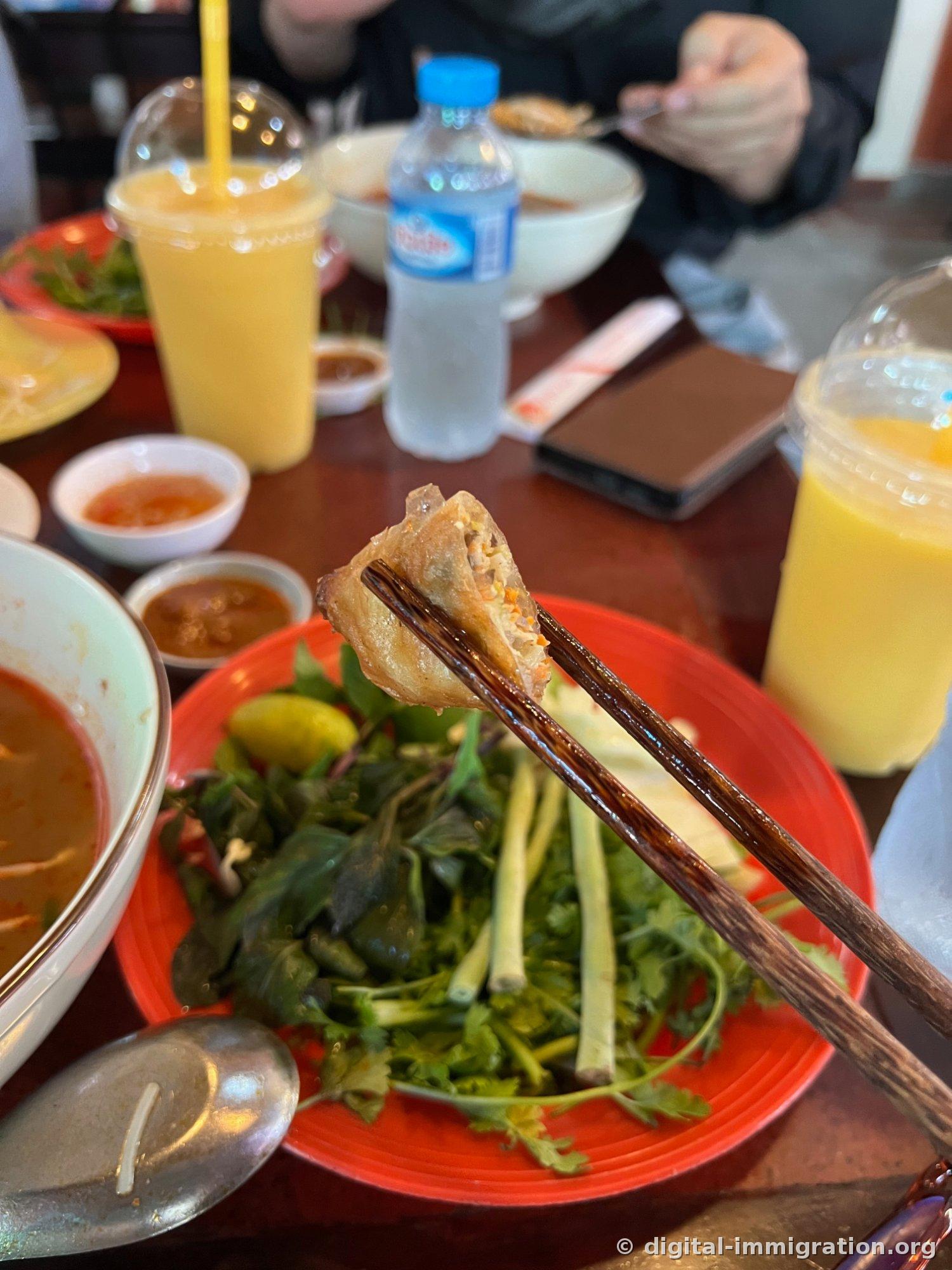 A crispy fried dumpling held with chopsticks, a red plate with basil, cilantro, water spinach, and lime, mango juice and water bottles with sauce dishes on the table