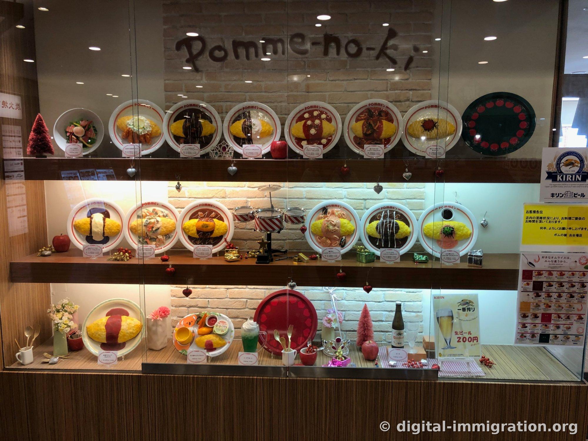 Un présentoir à trois niveaux sur un mur en briques avec une signature manuscrite 'pomme-no-ki'. Divers modèles d'omurice avec différentes sauces et des ornements de Noël sont exposés