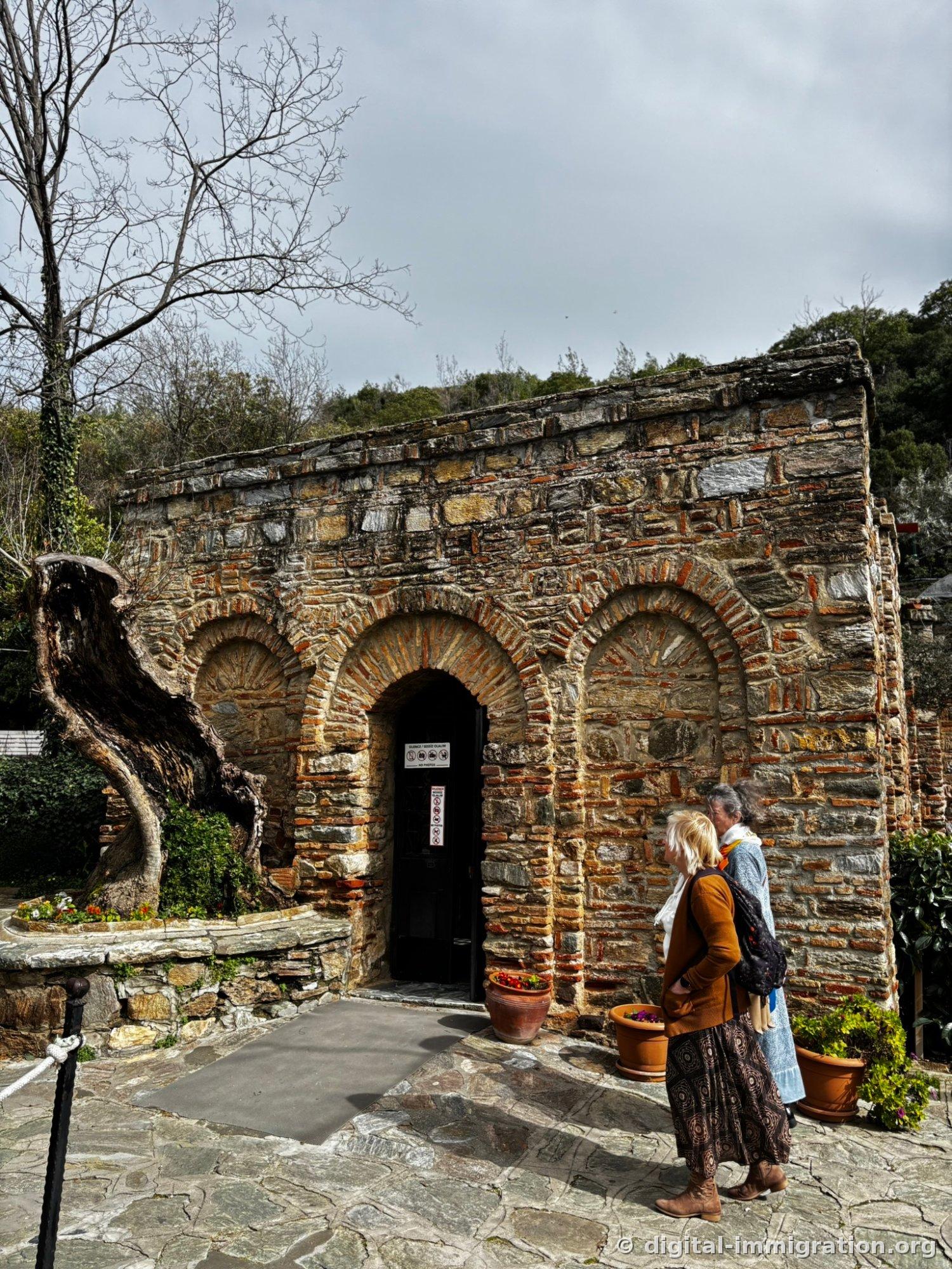 Exterior of the House of the Virgin Mary, a small three-arched building built of stone and red brick with the center arch serving as the entrance and a hollow old tree stump on the left