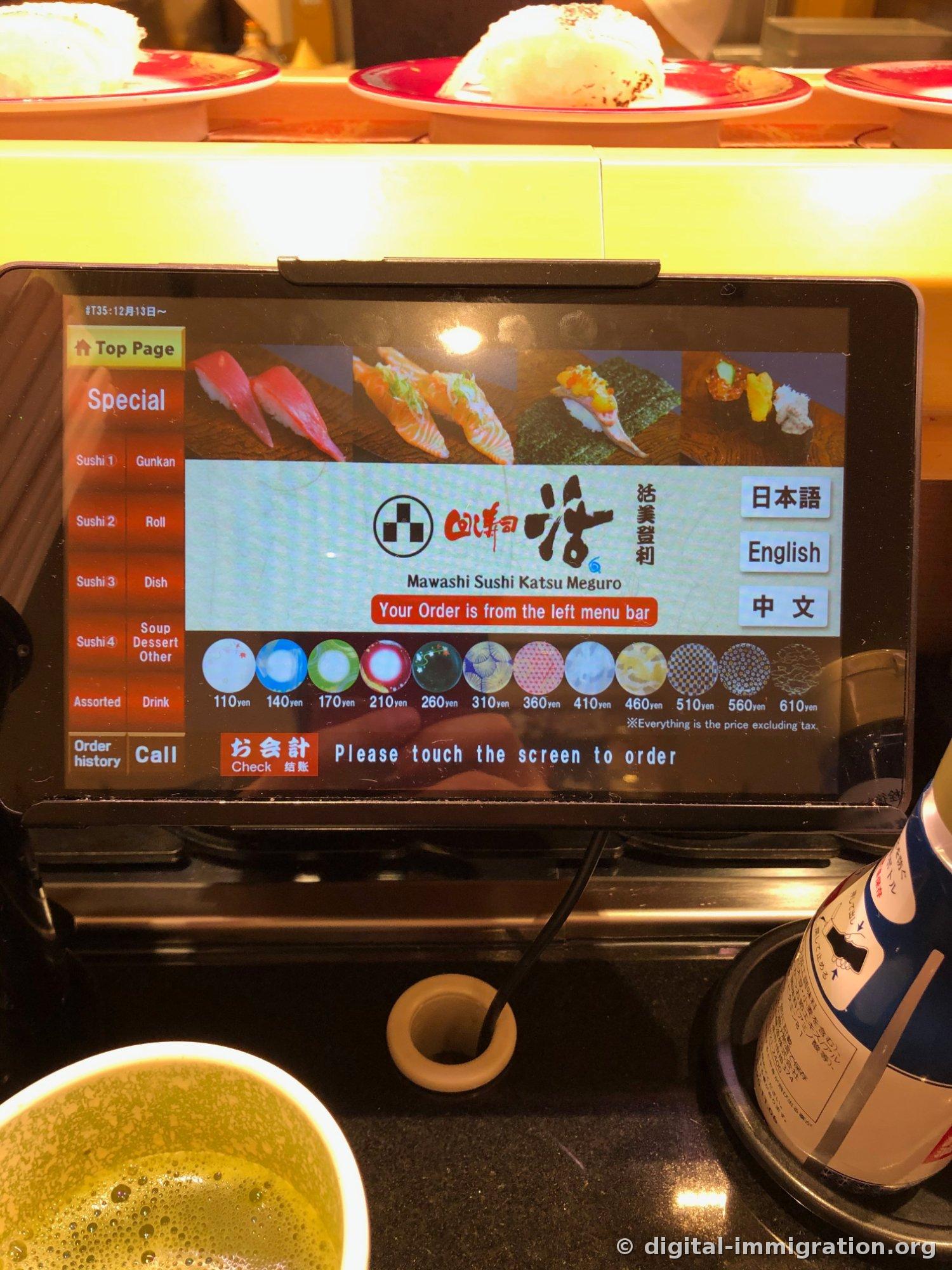吧台上放着的点餐平板屏幕，显示「Mawashi Sushi Katsu Meguro」标志和110日元至610日元的盘子颜色价格表