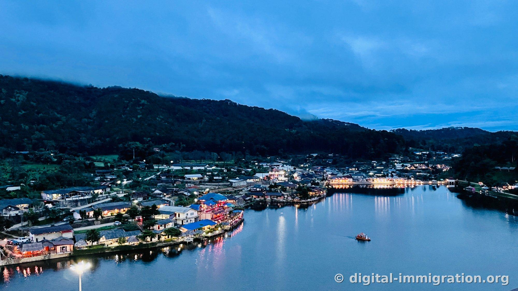 Vue d'ensemble du lac et du village Ban Rak Thai au crépuscule depuis une hauteur. Les lumières des commerces en bordure de lac se reflètent longuement dans l'eau, et une petite barque flotte à droite