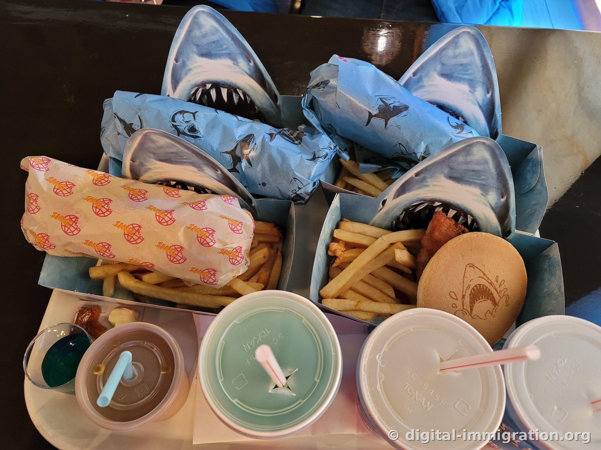 French fries in a shark-mouth-shaped paper box, sandwich wrapped in blue shark-patterned paper, burger bun with shark branding iron mark, and various drink cups