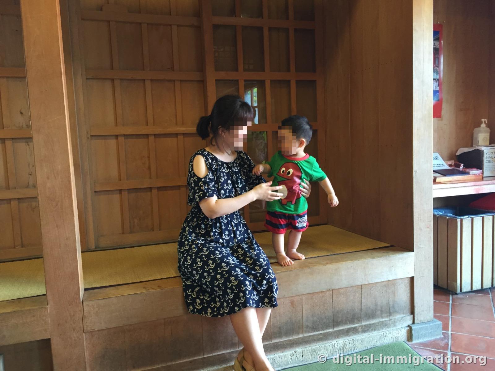 An adult sitting on tatami in a rest area with wooden pillars and a barefoot child standing beside, reception desk visible inside