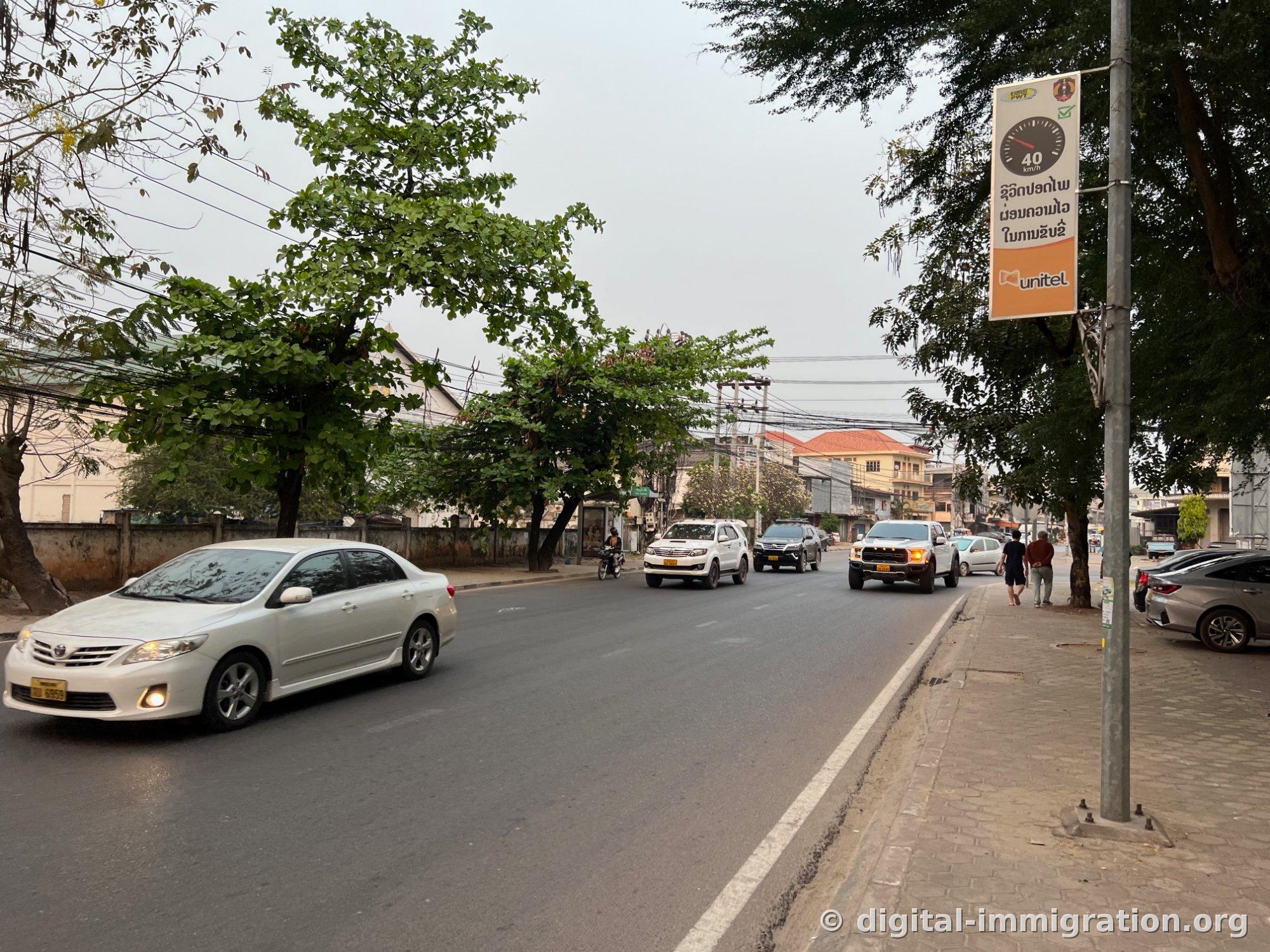 A street in central Vientiane. A white Toyota sedan and pickup truck pass by, while a power line on the right displays a 40km/h speed limit banner with text in Lao and a Unitel advertisement