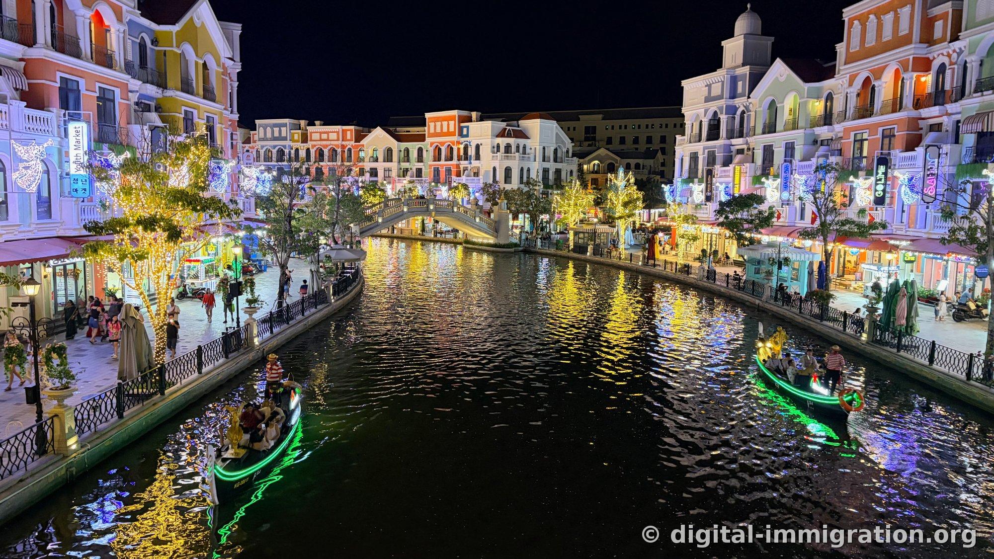 Night canal with illuminated buildings on both sides and yellow light reflections stretching across the water, two gondolas with green neon lights passing by