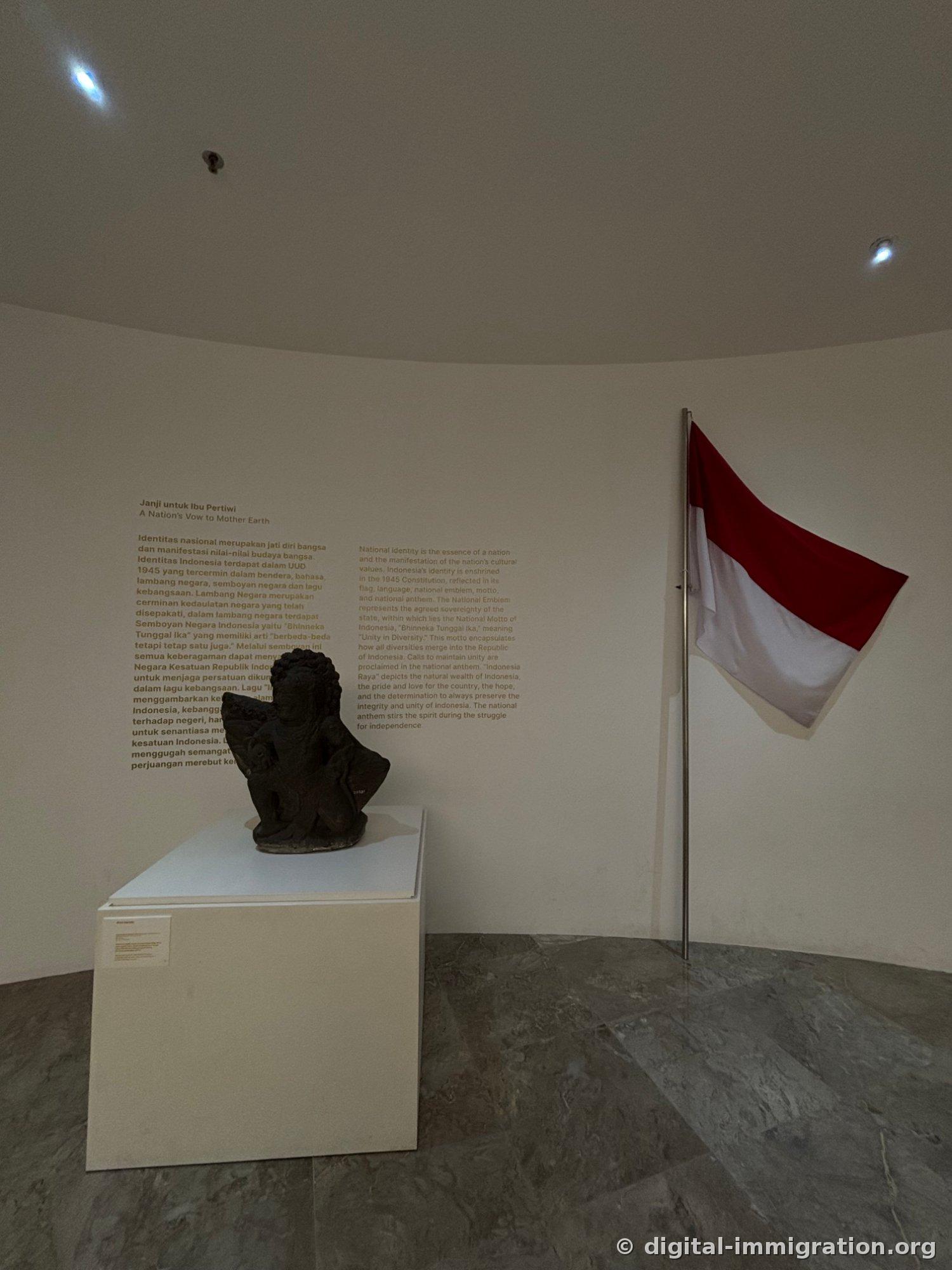 A dark circular exhibition room with 'Janji untuk Ibu Pertiwi' text on a curved wall, a white pedestal with a statue, and the Indonesian flag draped on the right