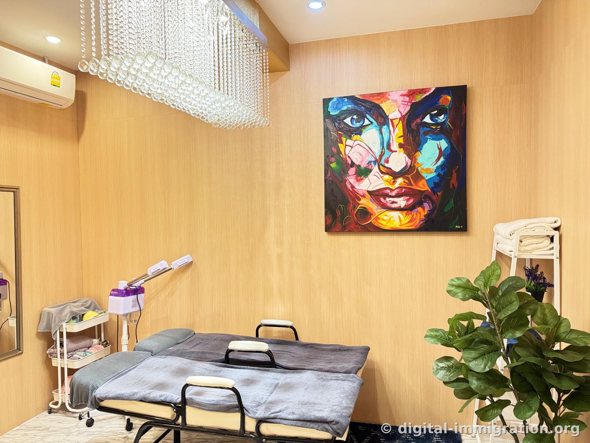 Interior of individual treatment room with facial bed covered in gray towel, steamer, magnifying lamp, brightly painted face painting on wall, glass bead lighting and wall-mounted air conditioner