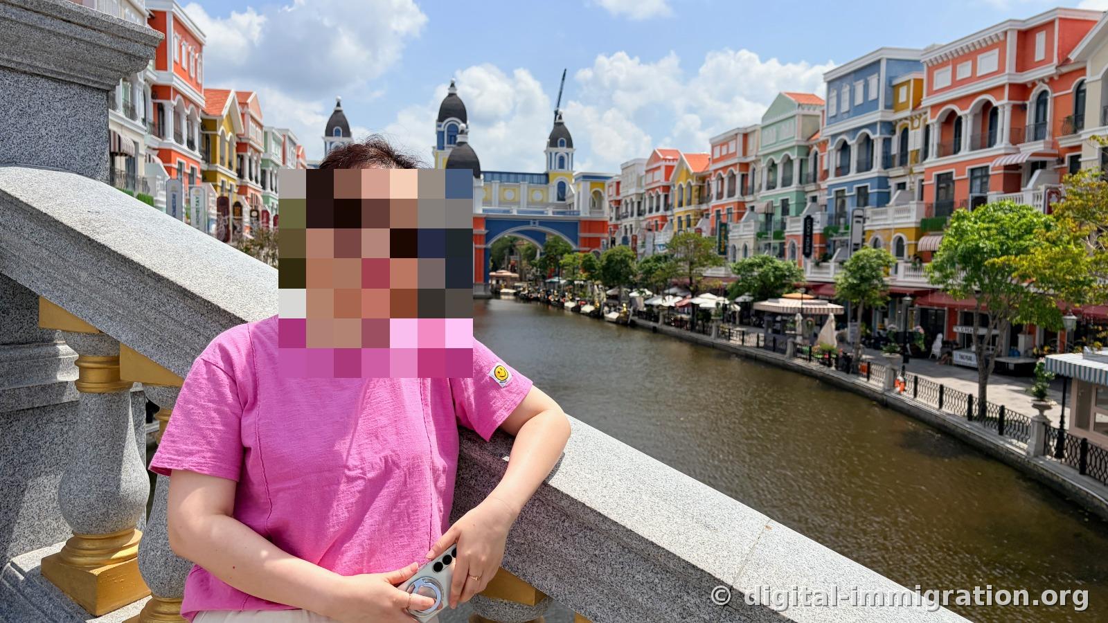 A person leaning against a stone bridge railing with colorful European-style buildings lining both sides of the canal behind them