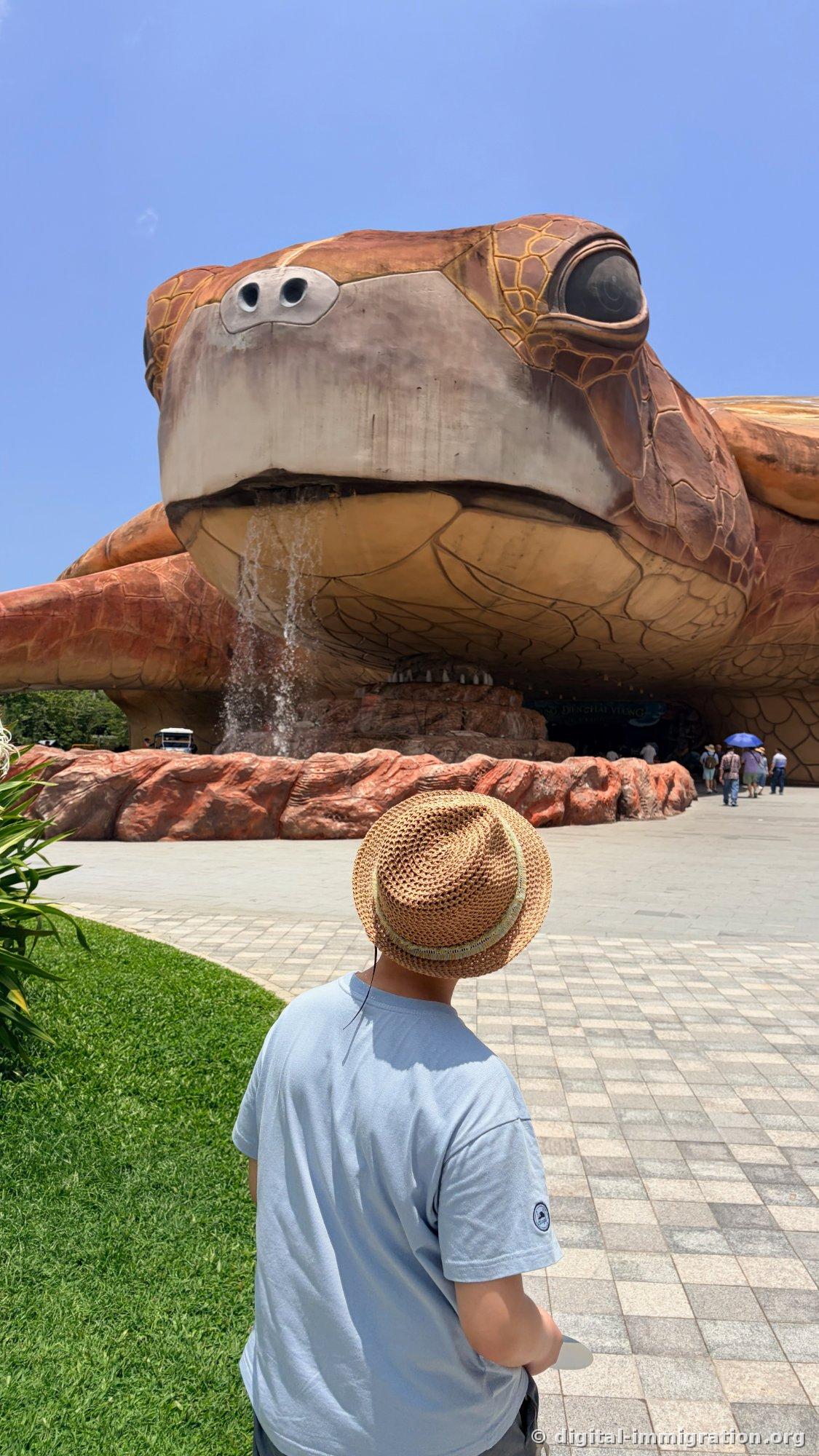A child in a straw hat looking up at the waterfall pouring from the turtle's head and mouth