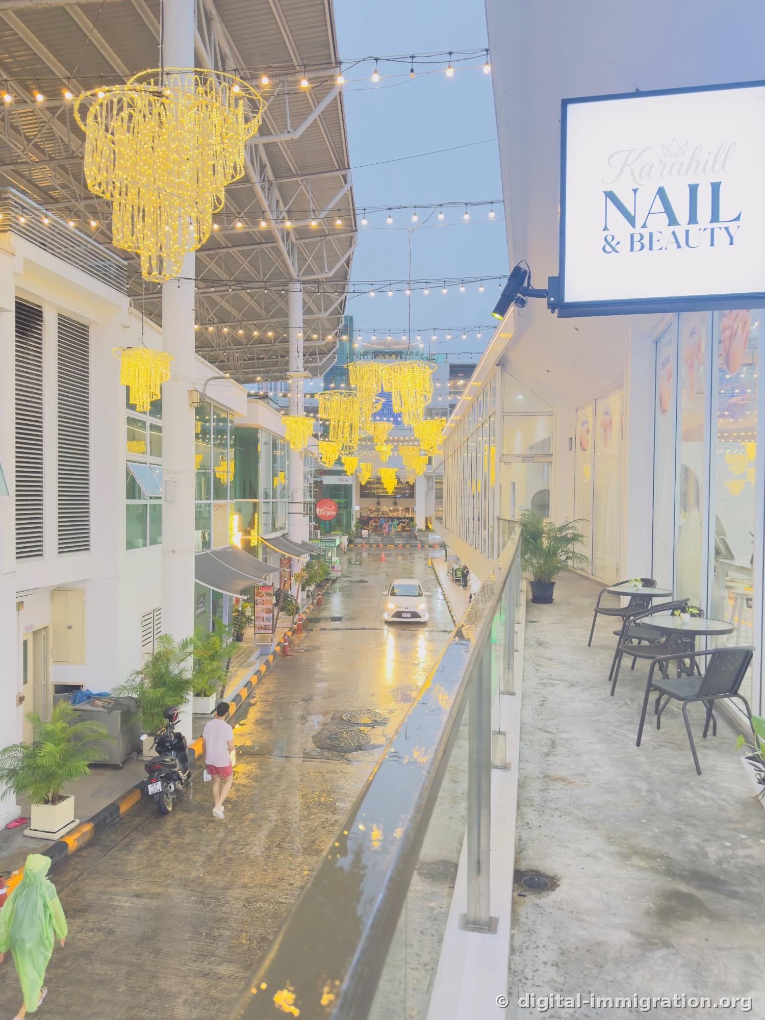 View down into the shopping alley at dusk from the 2nd floor railing, yellow chandeliers and light strands glowing in the corridor, wet pavement reflecting the lights, Karahill NAIL & BEAUTY sign visible on right