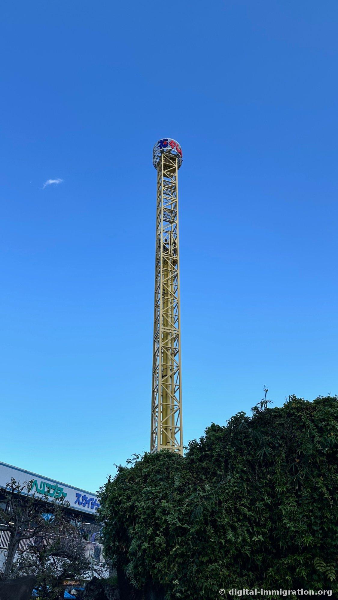 Yellow steel tower Space Shot ride extending to blue sky, with helicopter and sky ship signs on blue building below