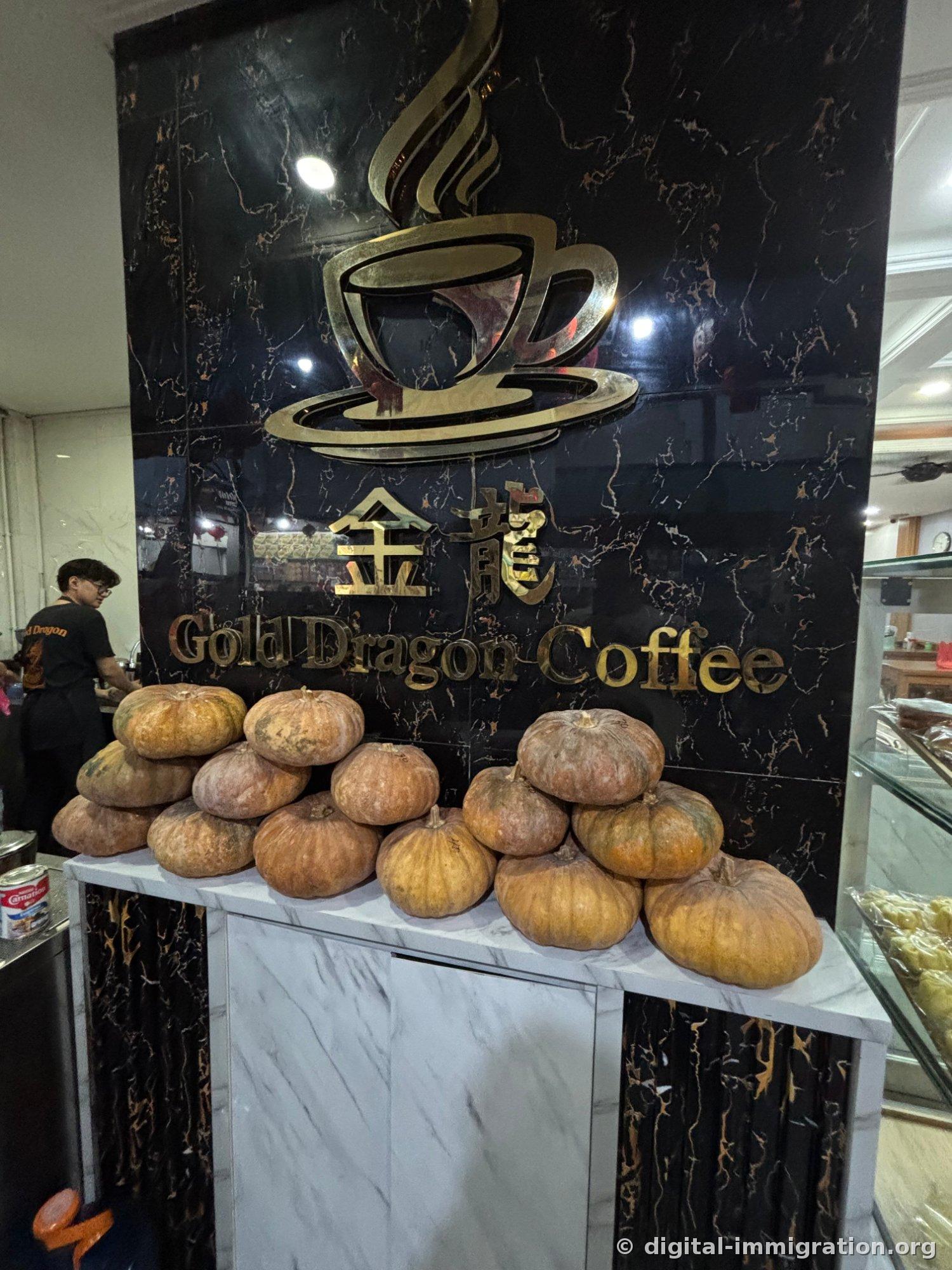 Logo de tasse de café doré et caractères 金龍 Gold Dragon Coffee sur un mur de marbre noir, avec plusieurs vieilles courges empilées sur le comptoir de la caisse devant