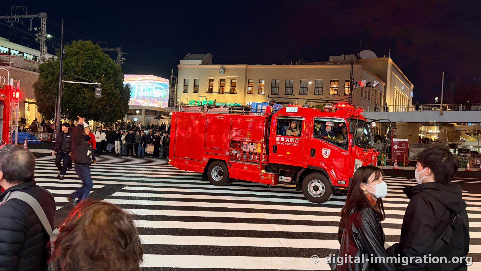 Un camion de pompiers rouge « 東京消防庁 TOKYO FIRE DEPT. » passant par le passage piéton devant la gare d'Ueno, des piétons le regardant, le bâtiment de la gare d'Ueno visible derrière