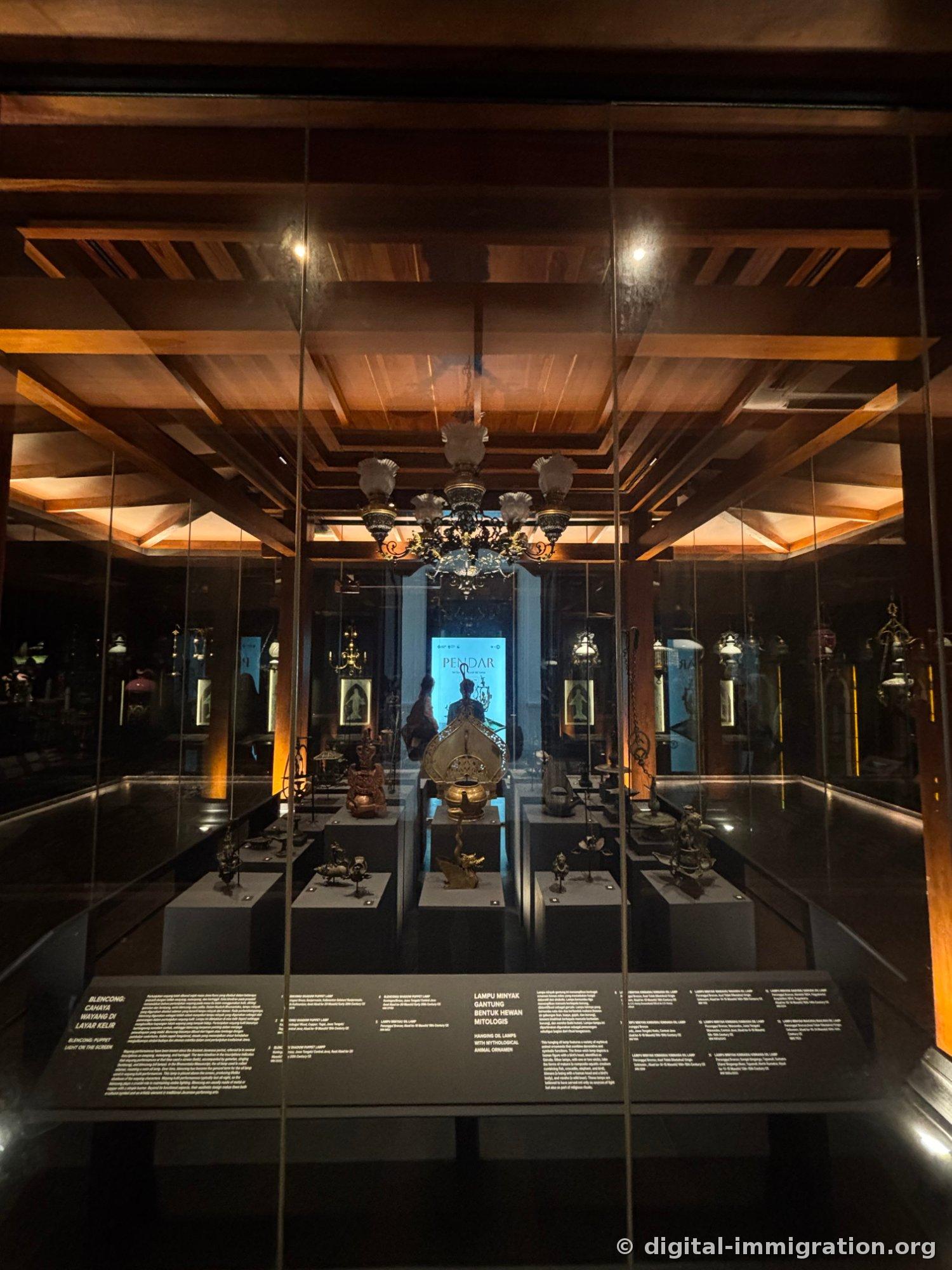 A dark exhibition room below a wooden ceiling with a hanging crystal chandelier, brass oil lamps on low pedestals, and a 'PENDAR' information panel