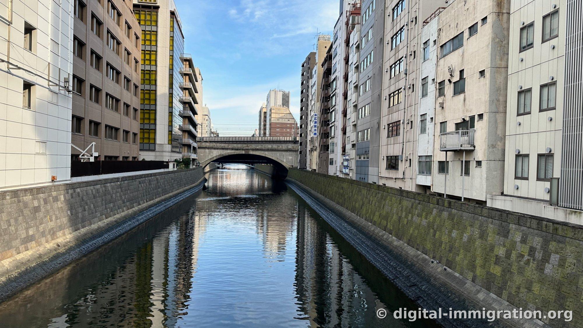 Clear morning scene on Kanda River with buildings and sky reflecting on calm water surface, arch bridge visible in distance