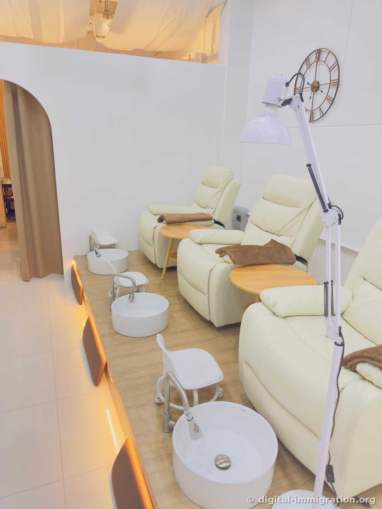 Three cream-colored pedicure recliners on wooden deck, white ceramic foot soak basins, wooden side tables, large white floor lamp and wall clock