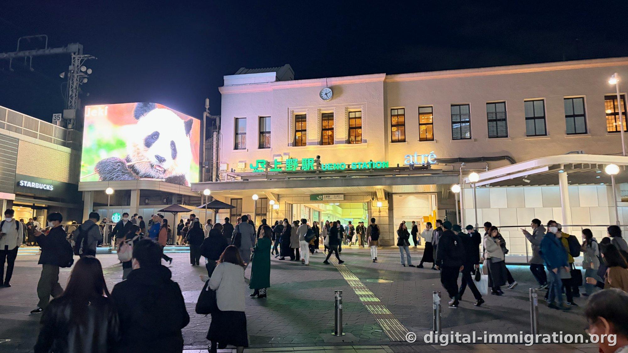 Le bâtiment de la gare JR Ueno illuminé la nuit avec l'enseigne « atré », un grand écran avec vidéo de panda à gauche, un Starbucks visible en bas, des gens circulant dans la place