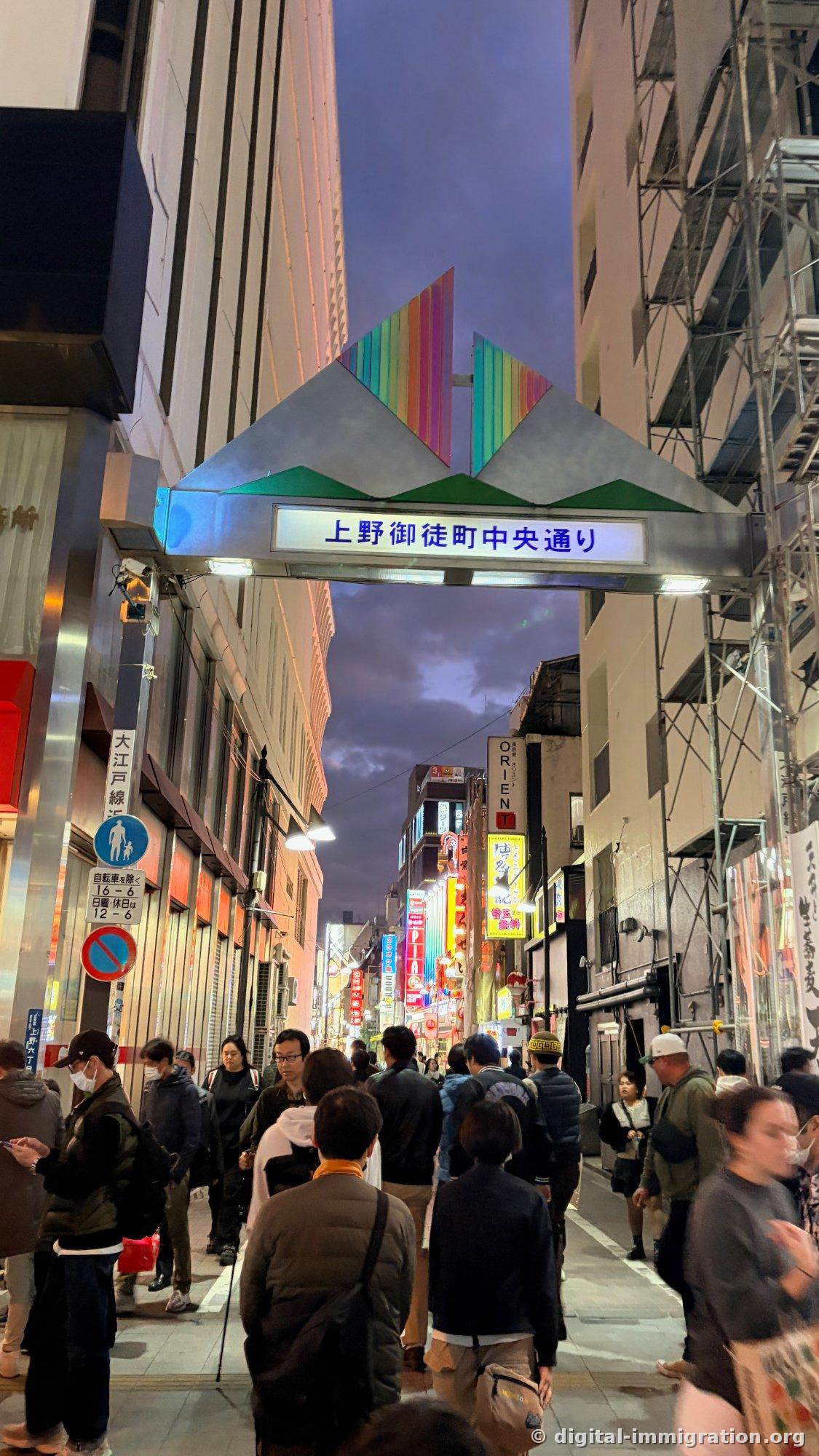 Un portail arc-en-ciel inscrit « 上野御徒町中央通り » sous le ciel du soir, avec une foule passant dessous et des enseignes au néon alignées à l'intérieur de la ruelle