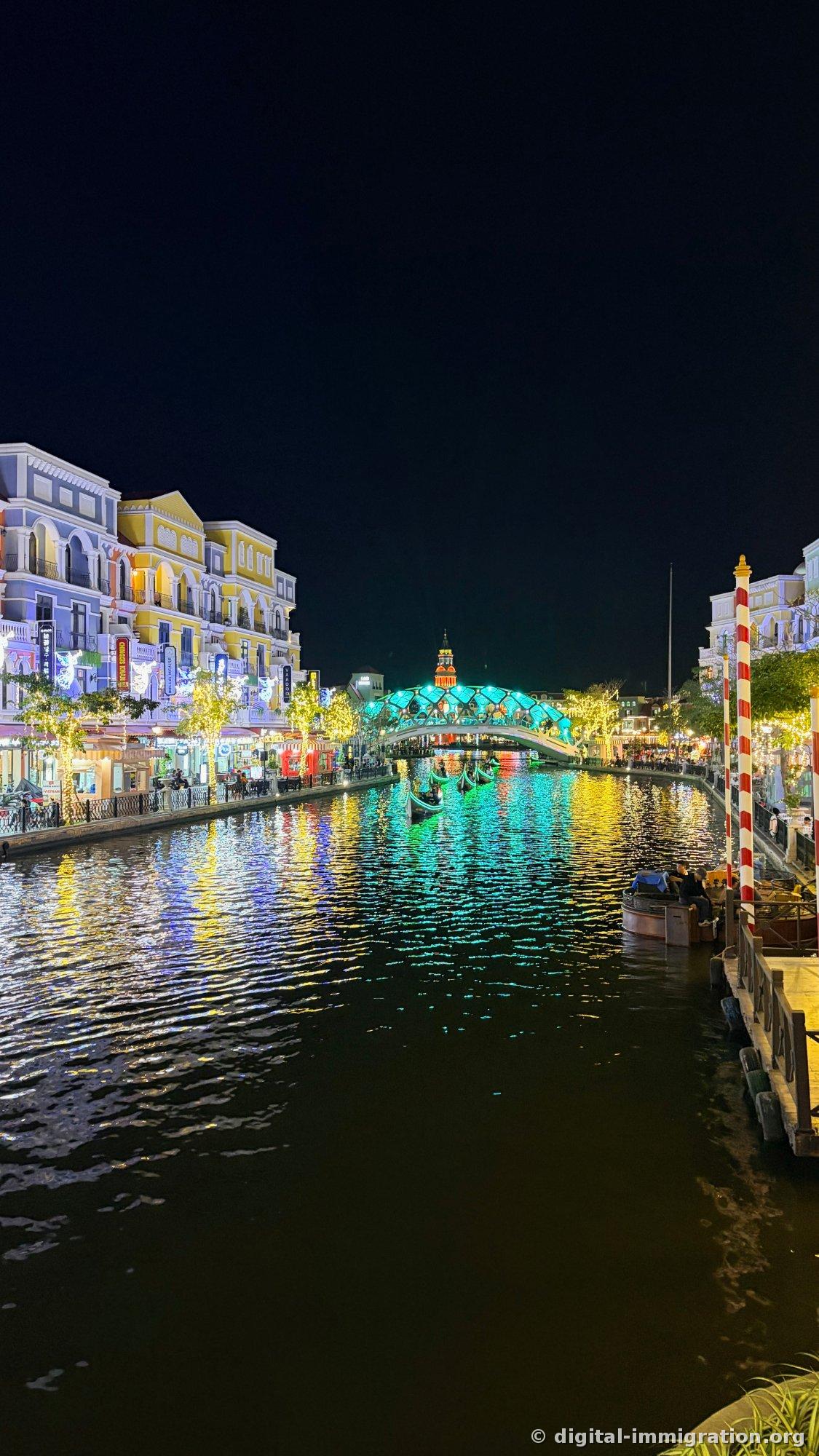 Vertical composition of the night canal with turquoise arch bridge and long yellow and blue light reflections stretching across the water