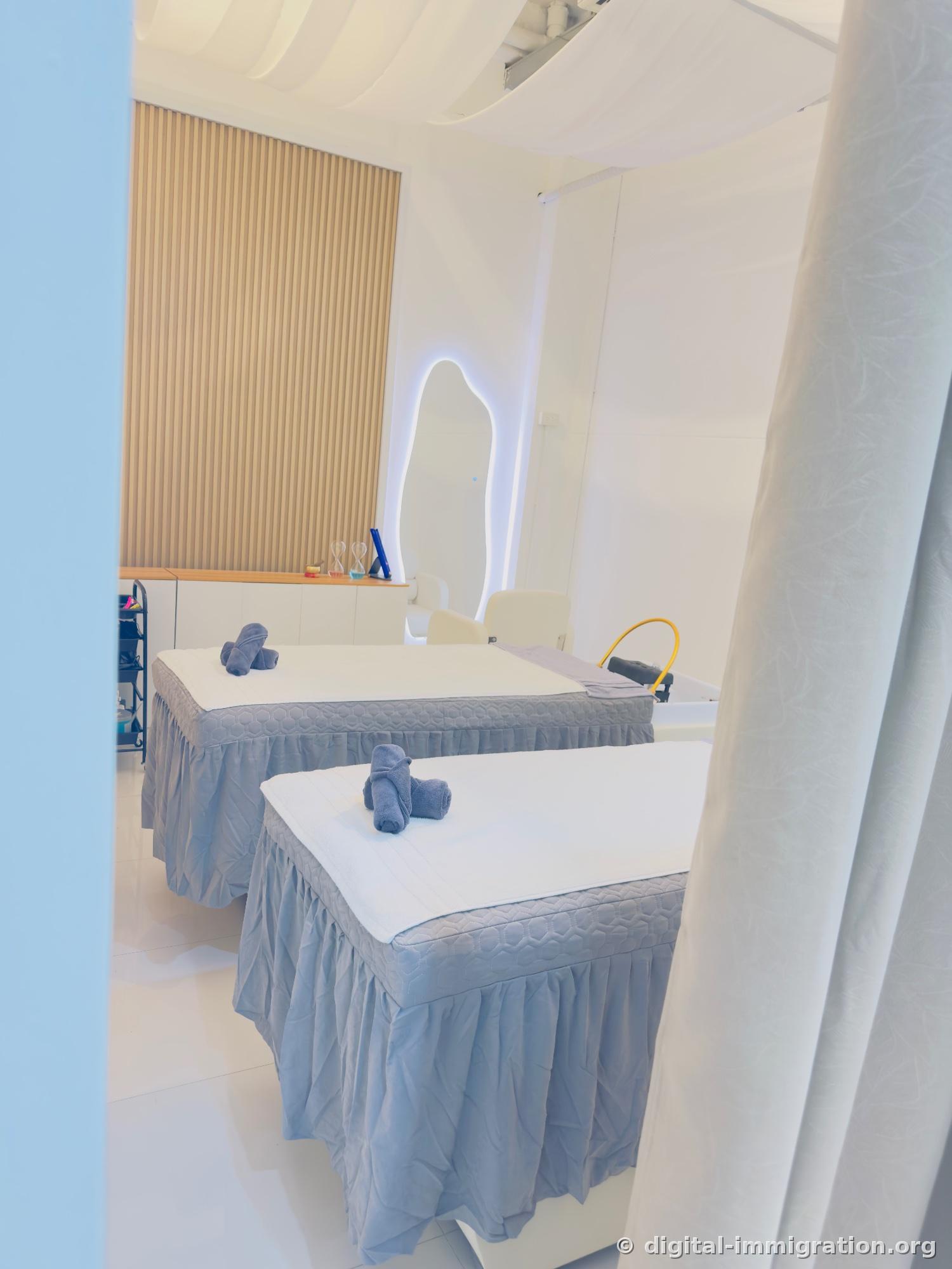 Treatment room beyond the white curtain, two beds with gray skirting with folded towels, wavy-edged illuminated mirror and head spa wash basin with yellow hose