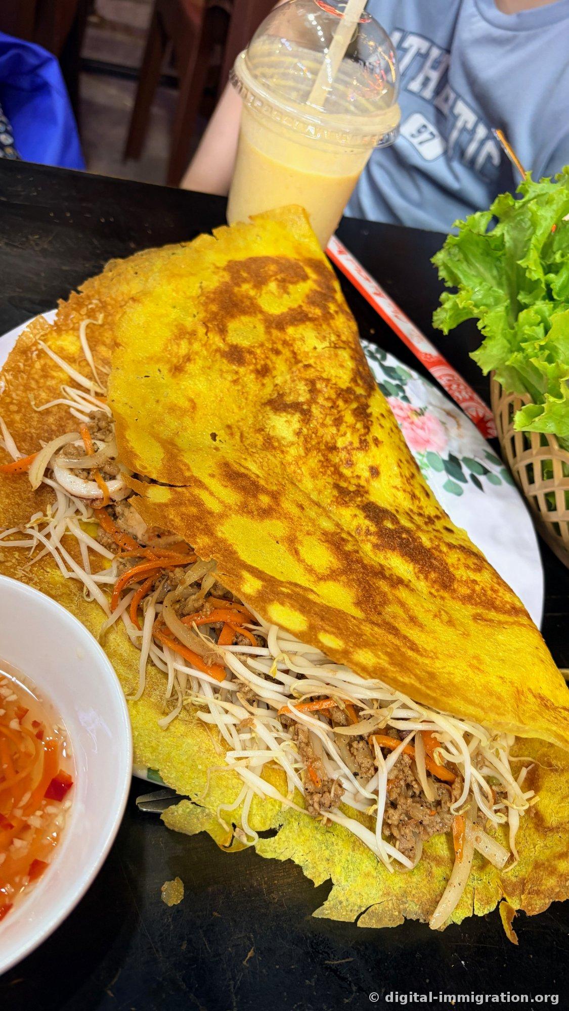 A large golden bánh xèo crepe filled with bean sprouts, shredded carrots and ground meat covering the plate, a basket of lettuce on the side, a mango smoothie in the back