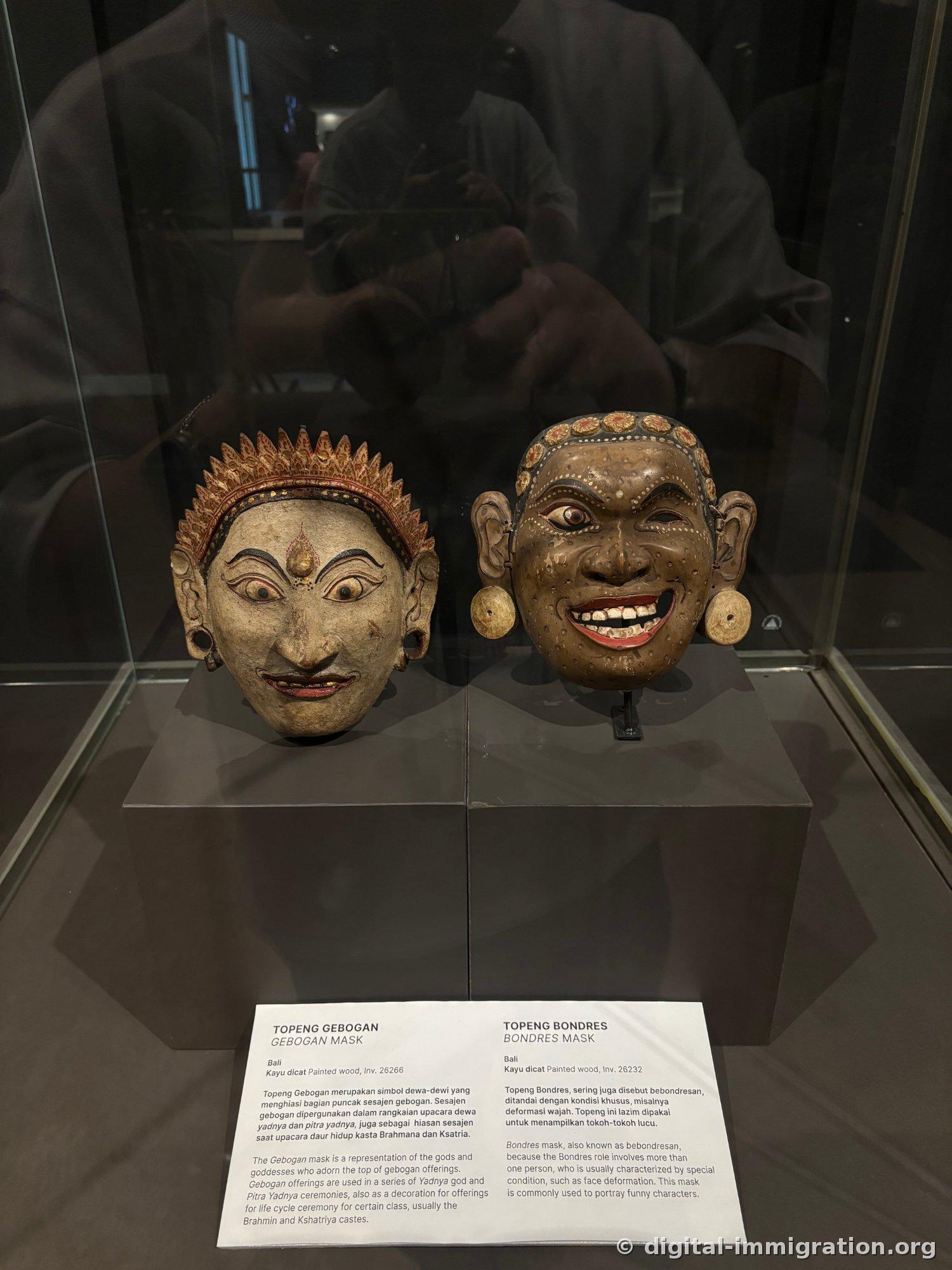 Two Balinese wooden masks side by side in a glass display case—the left with a white face and flame-shaped crown, the right with a brown face showing teeth and smiling