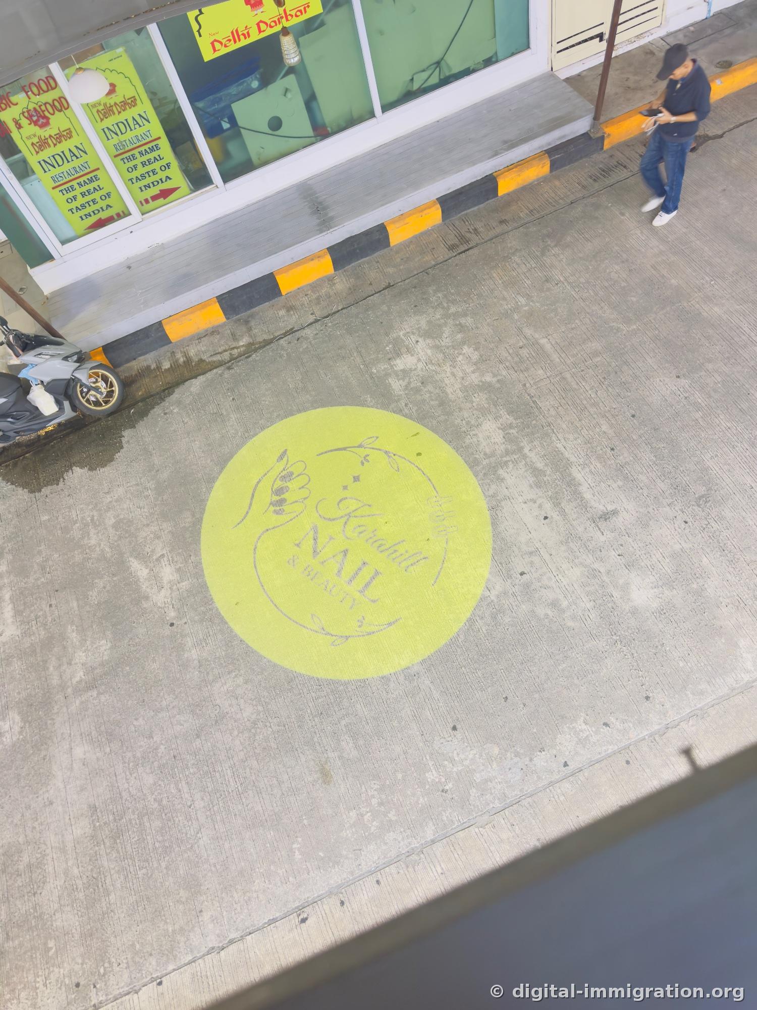 View of the 1st floor sidewalk from the 2nd floor, yellow circular Karahill Nail & Beauty logo painted on pavement and New Delhi Darbar Indian restaurant sign, motorcycles parked