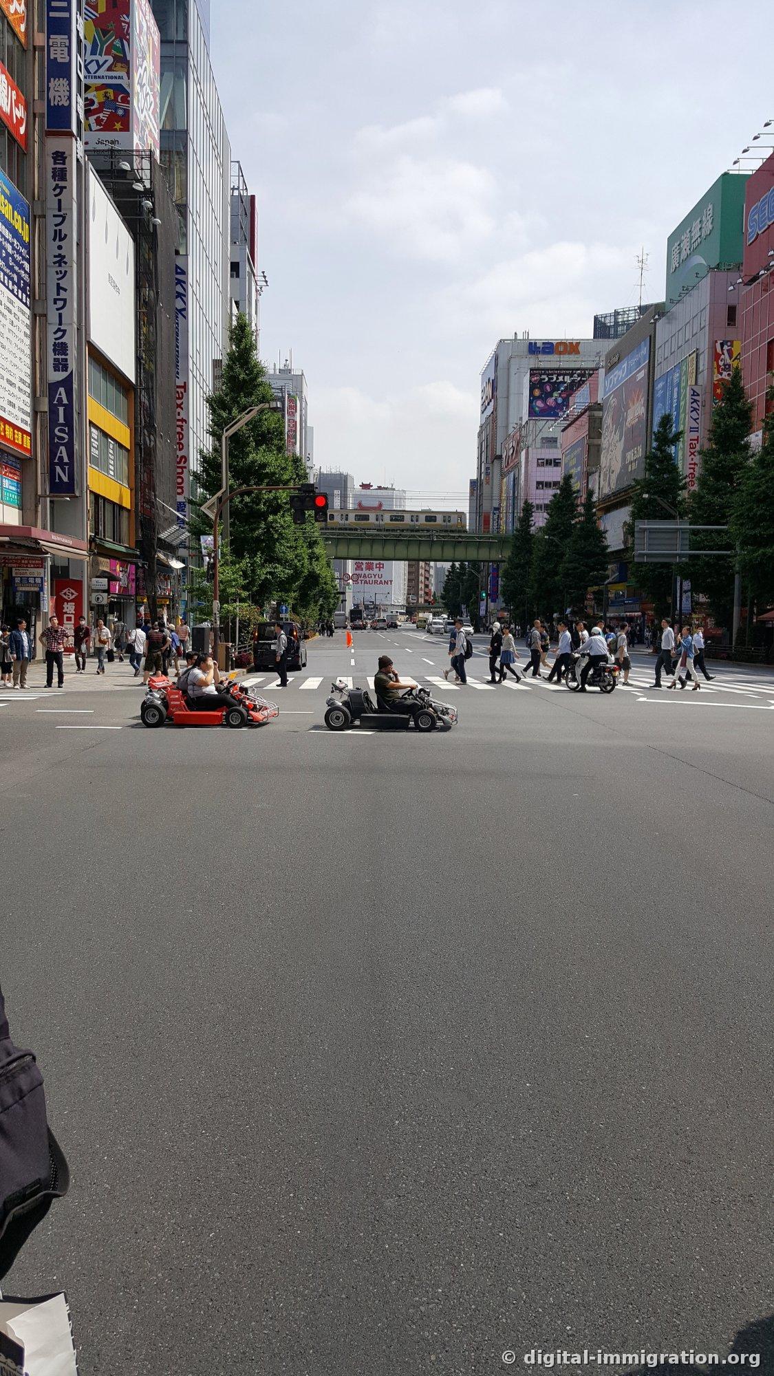 Two public road carts waiting for a traffic light on Chuo-dori, with the yellow Sobu Line train passing overhead on the elevated track