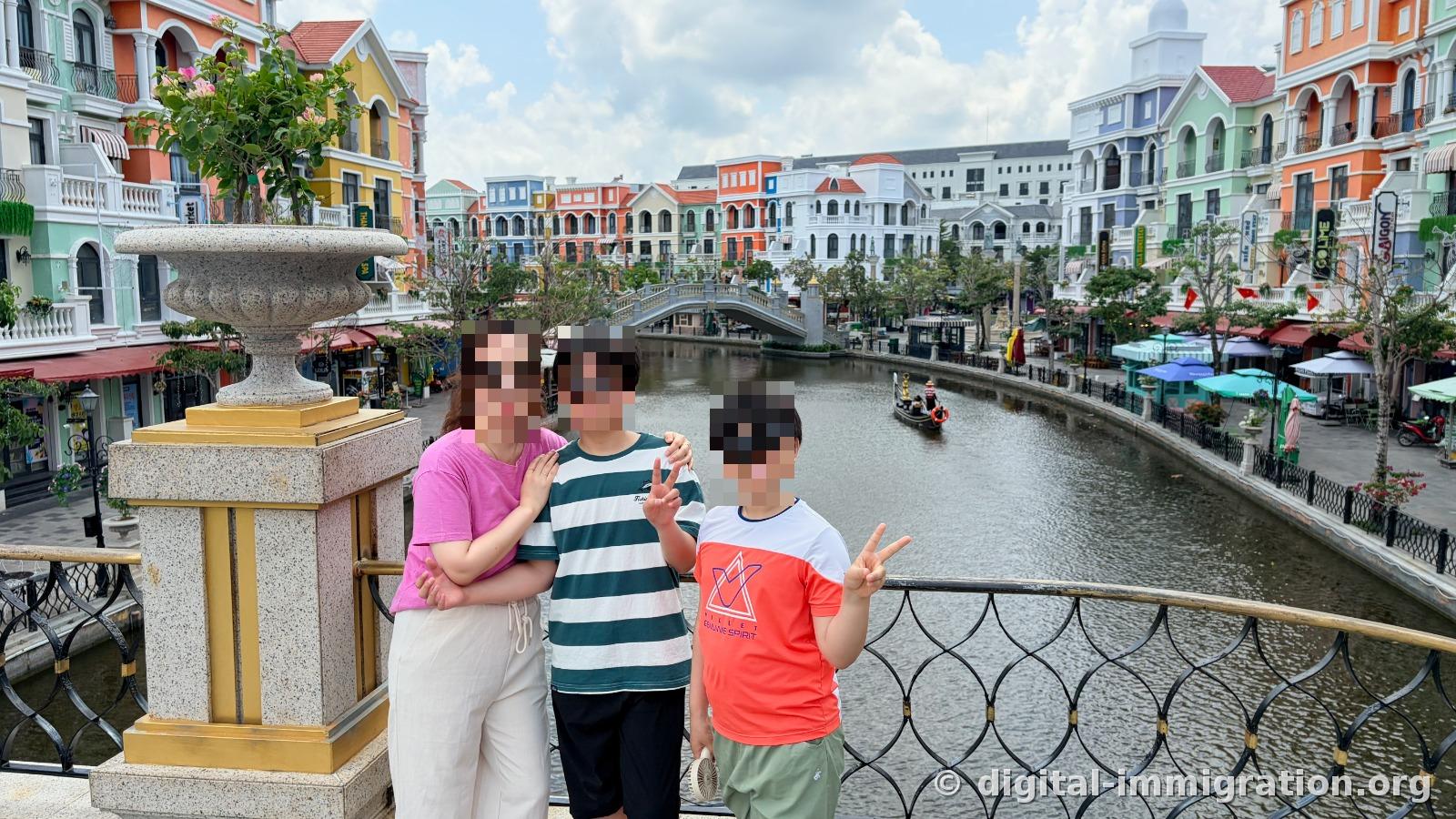 Three people standing together next to a stone planter on a bridge with a canal stretching diagonally behind them and a gondola in the distance
