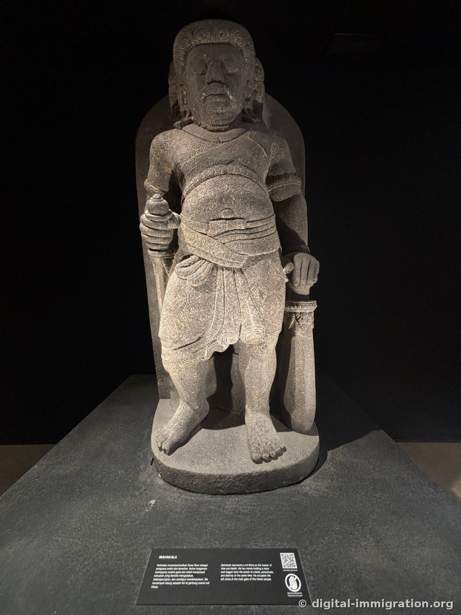The Mahakala stone statue illuminated against a dark background, with a protruding belly holding a club and a dagger
