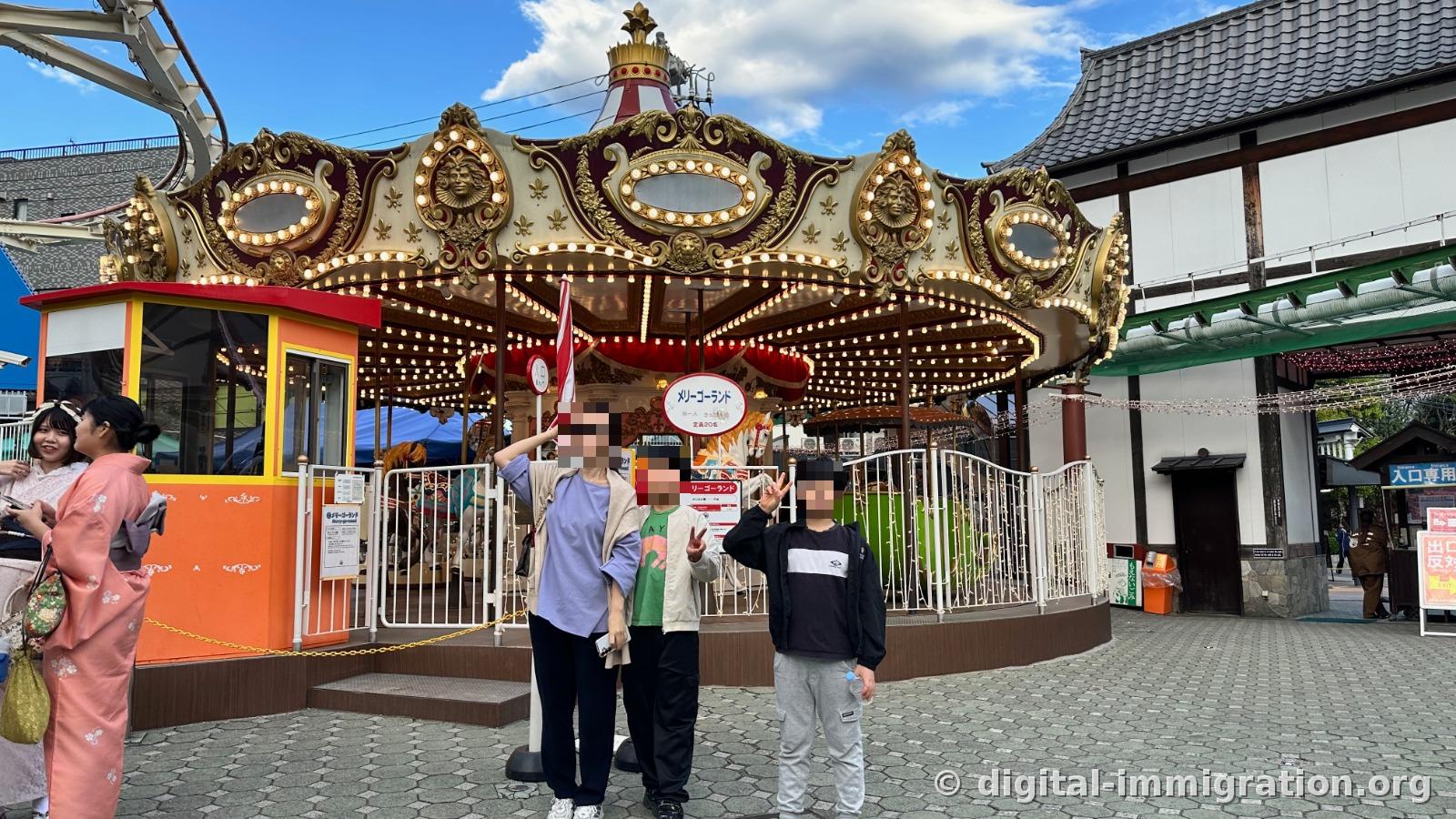 Three people posing in front of carousel covered in gold decorations and lights, with a tiled roof wooden building on the right