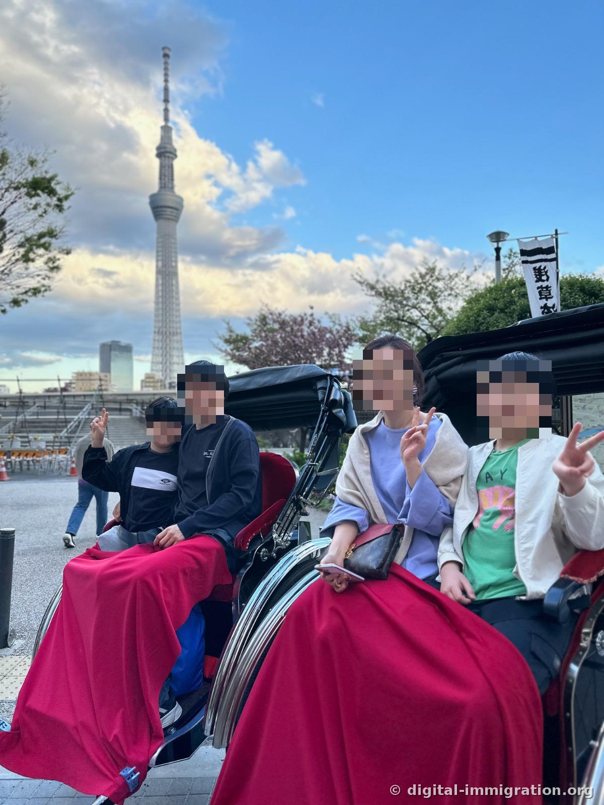 Four people split between two black rickshaws, covered with red lap blankets, striking peace signs with Skytree in the background.