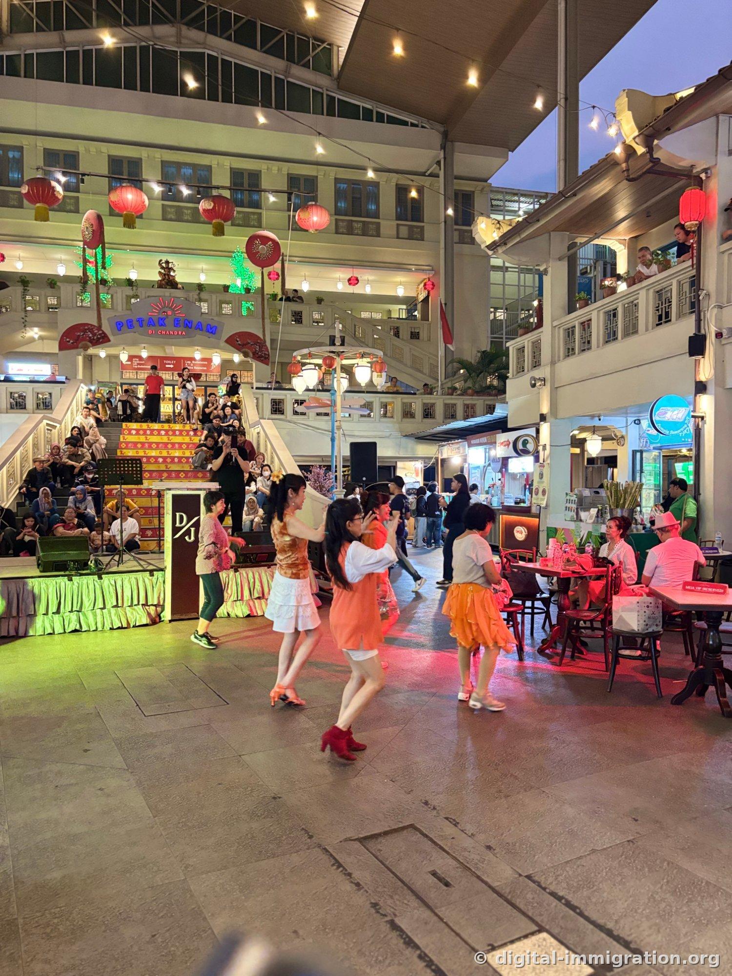 Women in orange and white clothing dancing in Petak Enam's central plaza, with people watching from yellow stepped bleachers and red lanterns hanging above