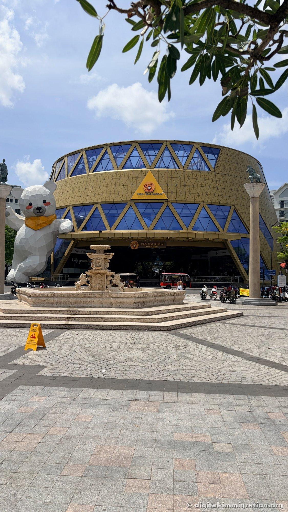 Round Teddy Bear Museum building with golden exterior and blue triangular glass windows, an angular white bear sculpture beside it, and a stone fountain in front