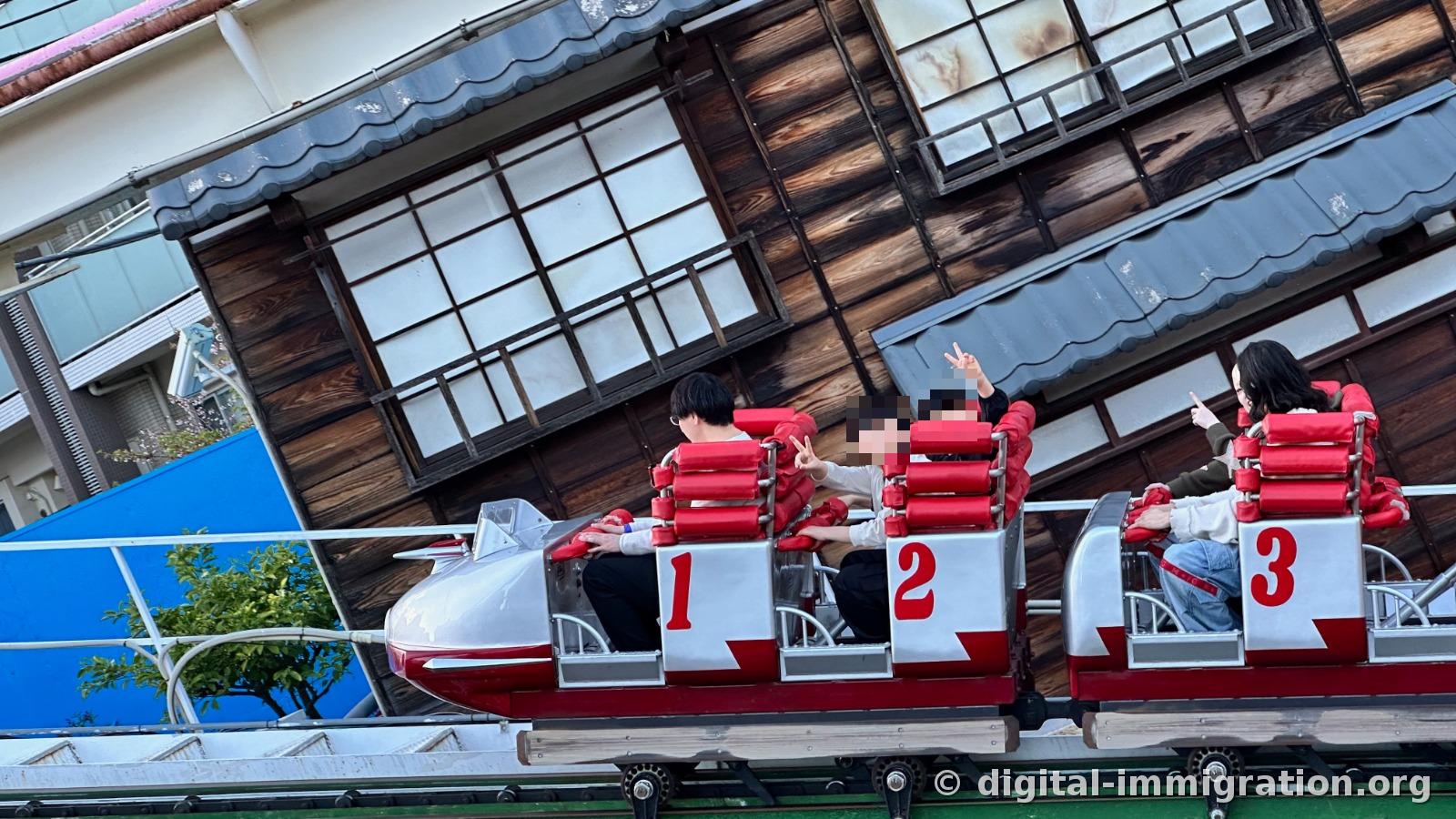 Red roller coaster car passing right next to the latticed window of an old wooden building, with passengers in seats 1, 2, 3 with hands raised