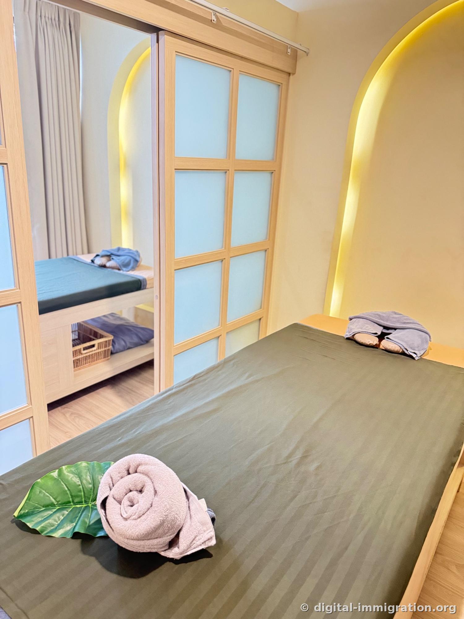 Massage room with arched yellow indirect lighting on walls, bed with khaki striped sheets, towels and artificial leaves on the bed, full-length mirror on one side and semi-transparent glass sliding door on the other