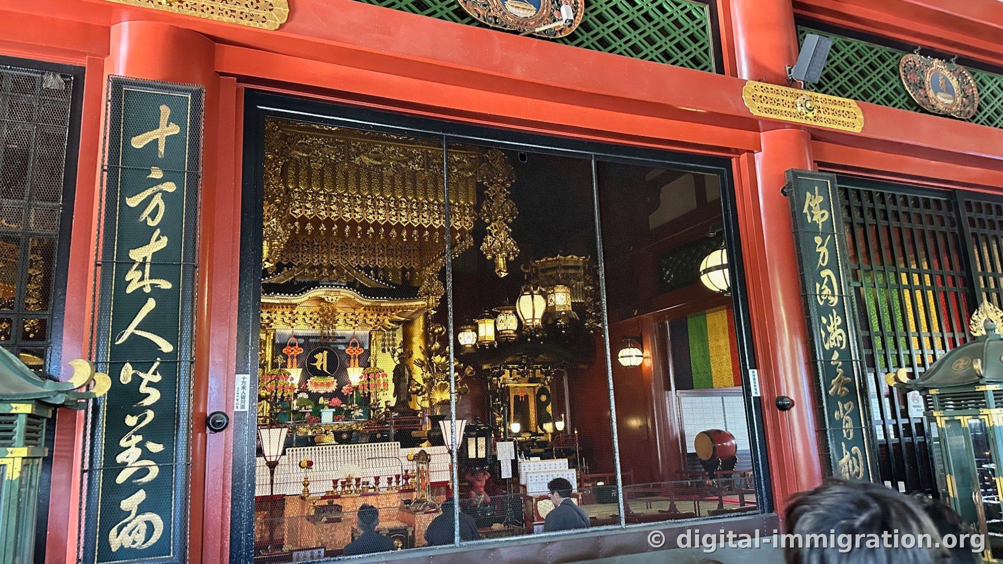 The interior of the Senso-ji main hall seen through the lattice, with golden canopies, lanterns, a red drum, and gold leaf inscription pillars on both sides