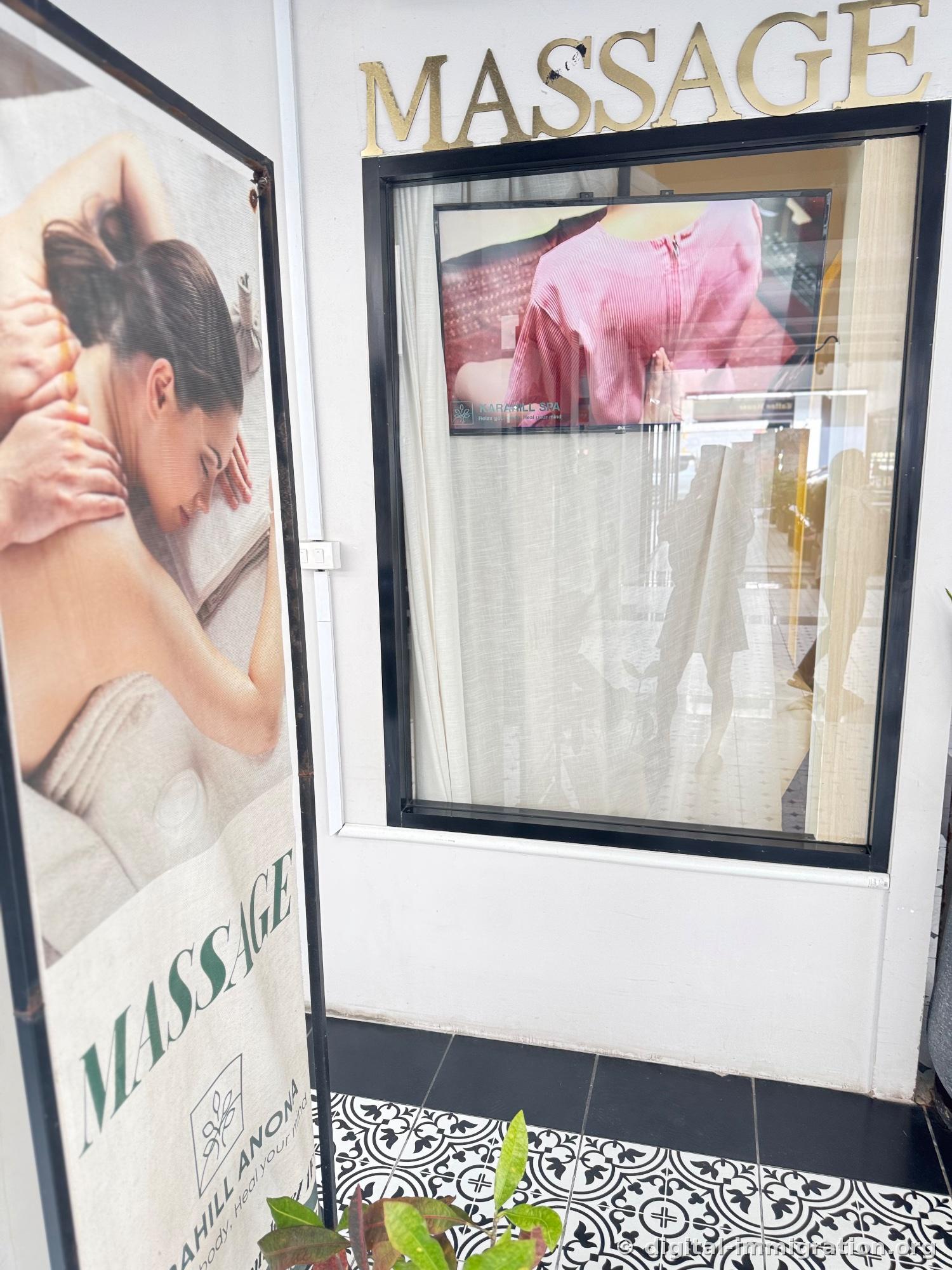Gold MASSAGE sign on white wall, window below showing promotional video on TV for karahill spa and massage photo banner, black-and-white patterned tile floor